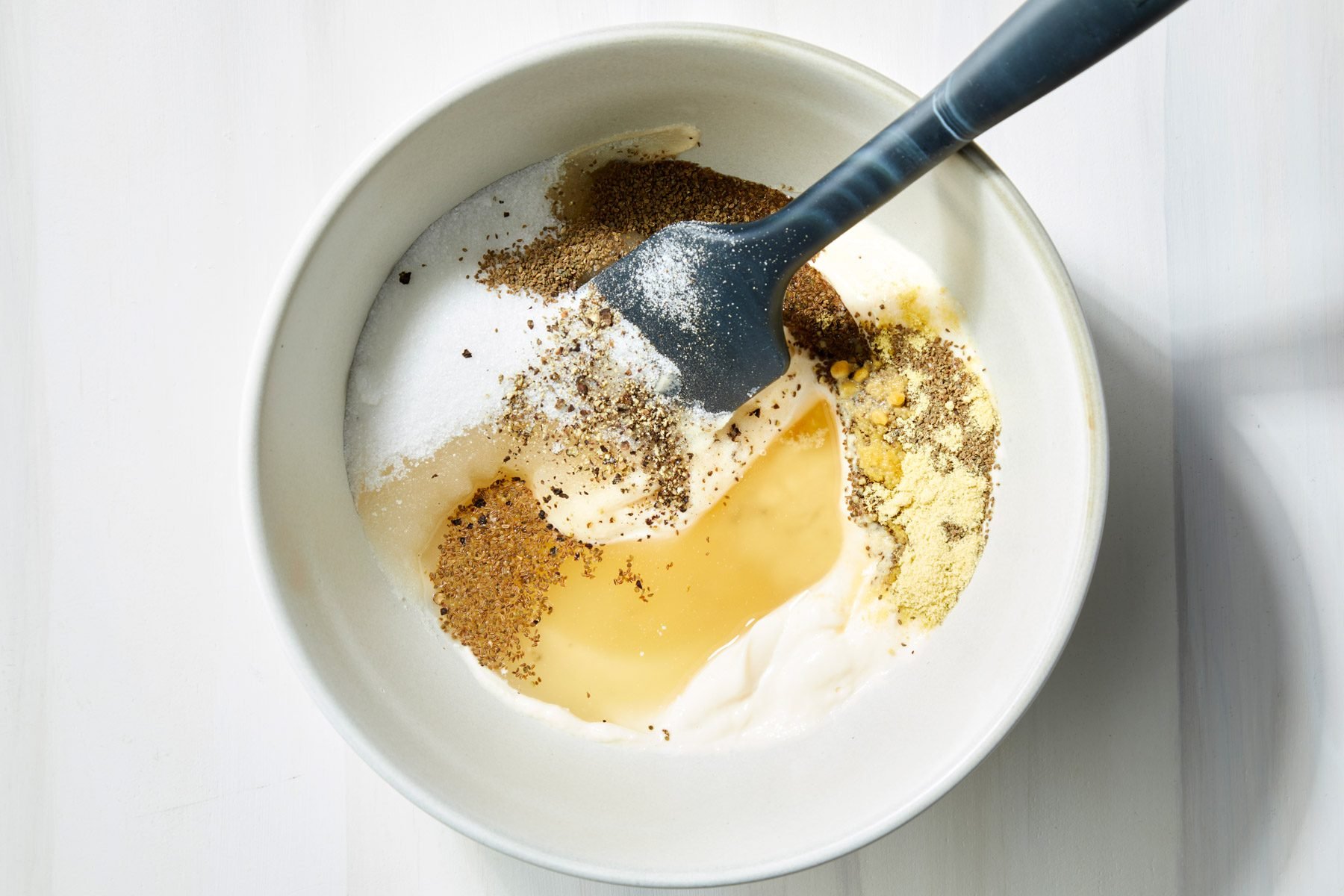 Mayonnaise, sugar, cider vinegar, celery seed, salt, pepper and ground mustard are being mixed in a white bowl with a spatula.