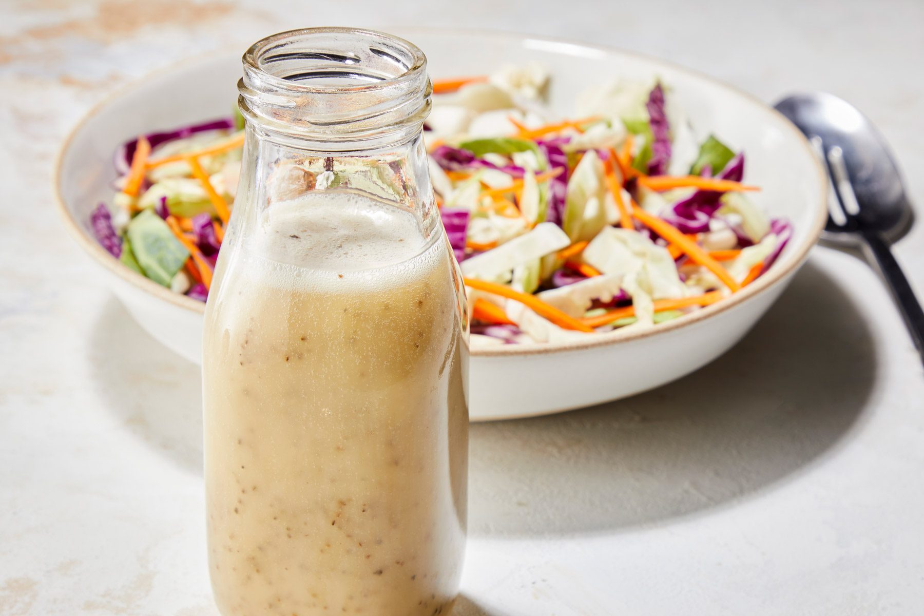 Coleslaw Dressing in front of a white bowl which contains salad. A silver spoon is next to the bowl.