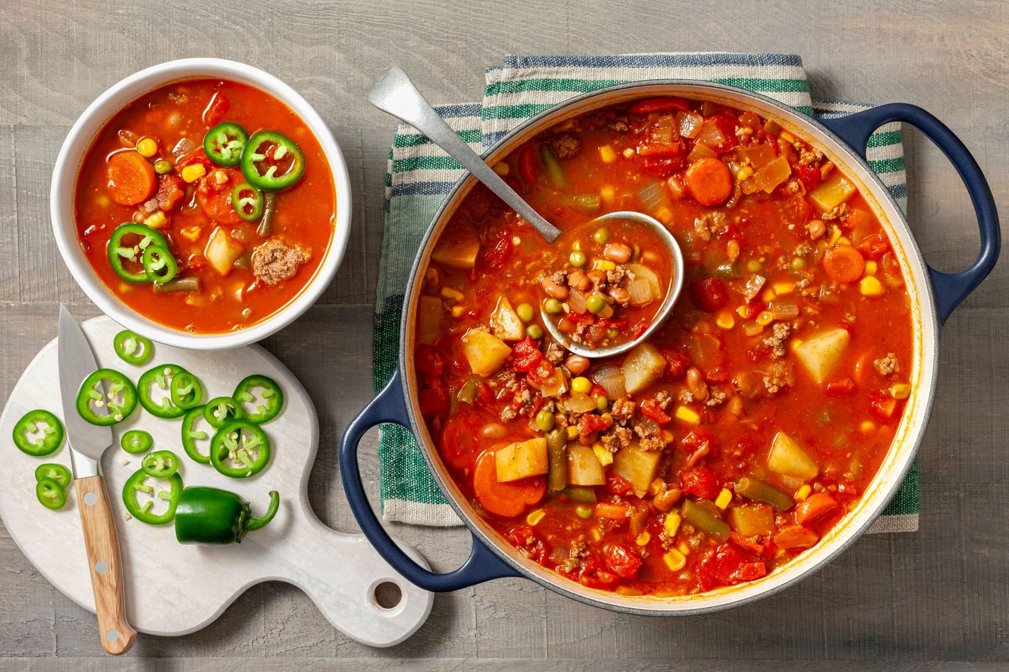 A large pot of cowboy soup with a ladle rests on a striped towel beside a white bowl filled with the same soup. Freshly sliced jalapeños and a knife are on a cutting board next to the bowl; wooden background;