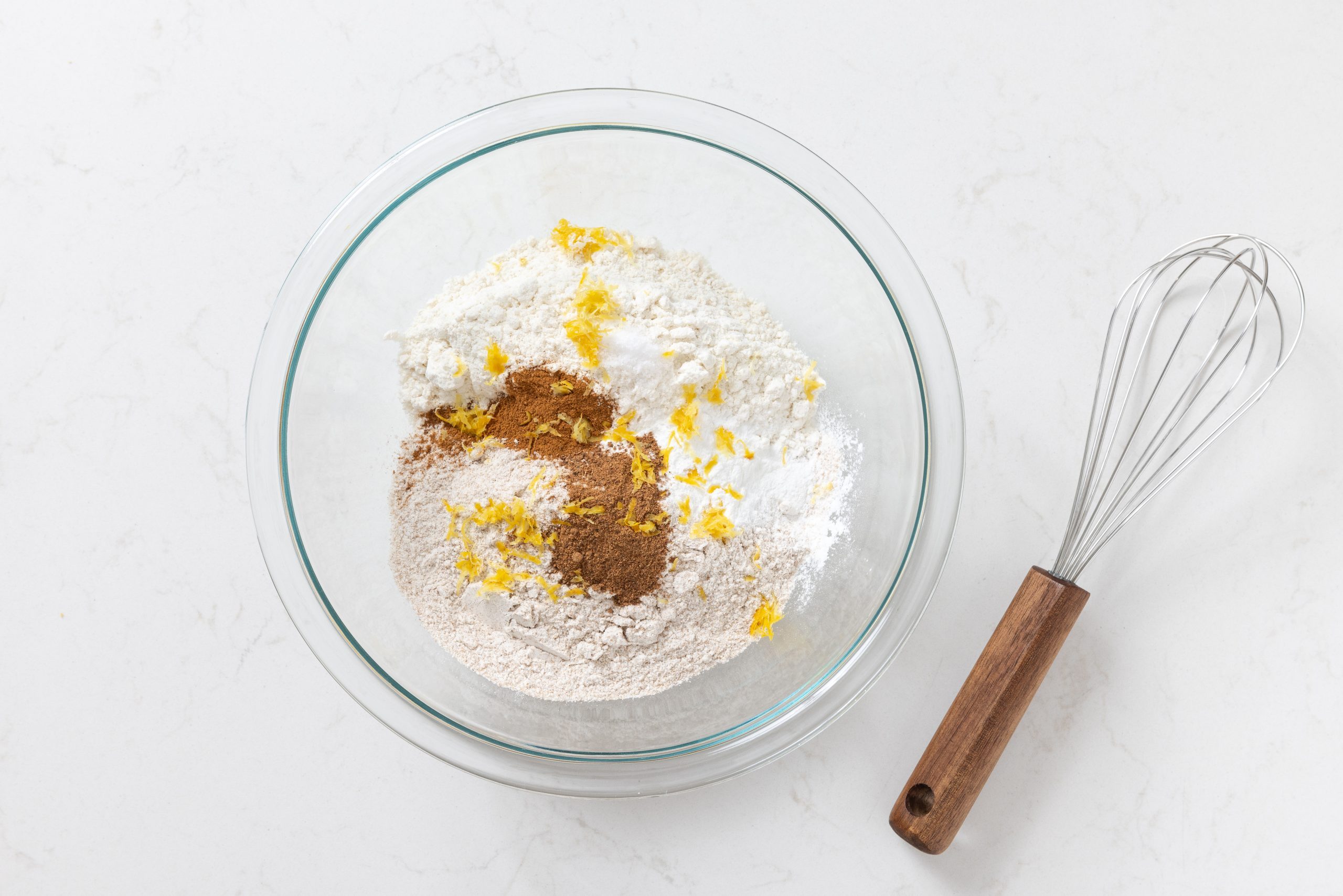 Flour, whole wheat flour, lemon zest, cinnamon, nutmeg, baking soda and salt being mixed in bowl.