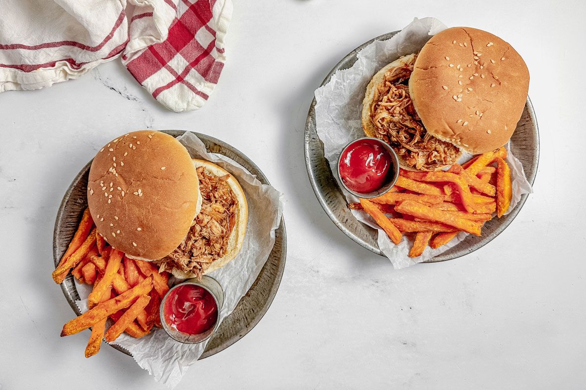 Taste of Home Dutch oven pulled pork on metal plates with sweet potato fries and tins of ketchup on a marble surface