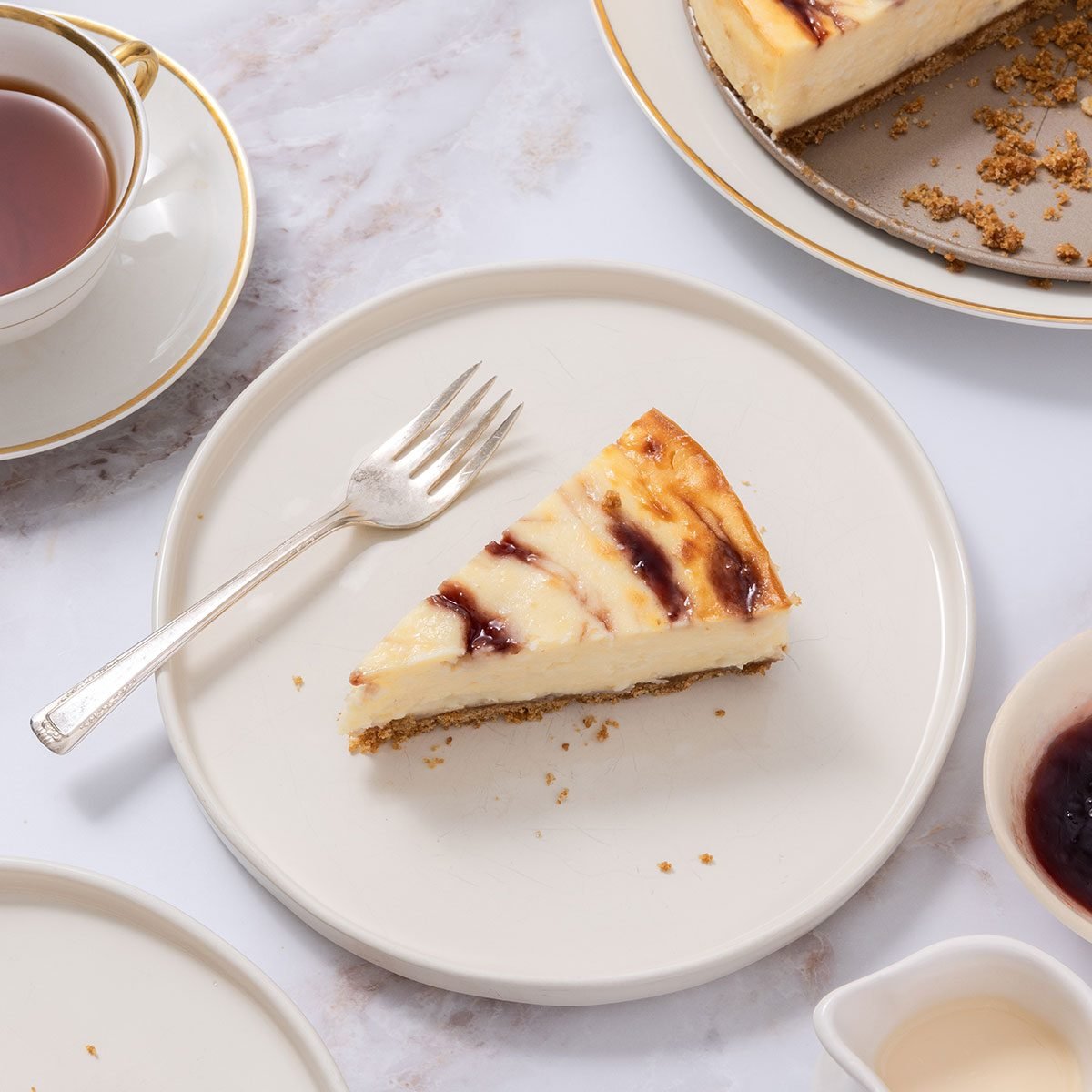 Cheesecake slice served on plate with full cheesecake near it, served with tea, cream and jam.