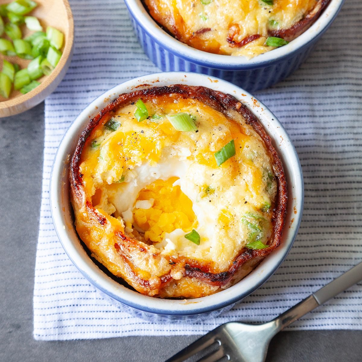 Taste of Home Bacon and Egg Muffins in blue ramekins, topped with green onions cheese, wooden bowl of green onions, forks