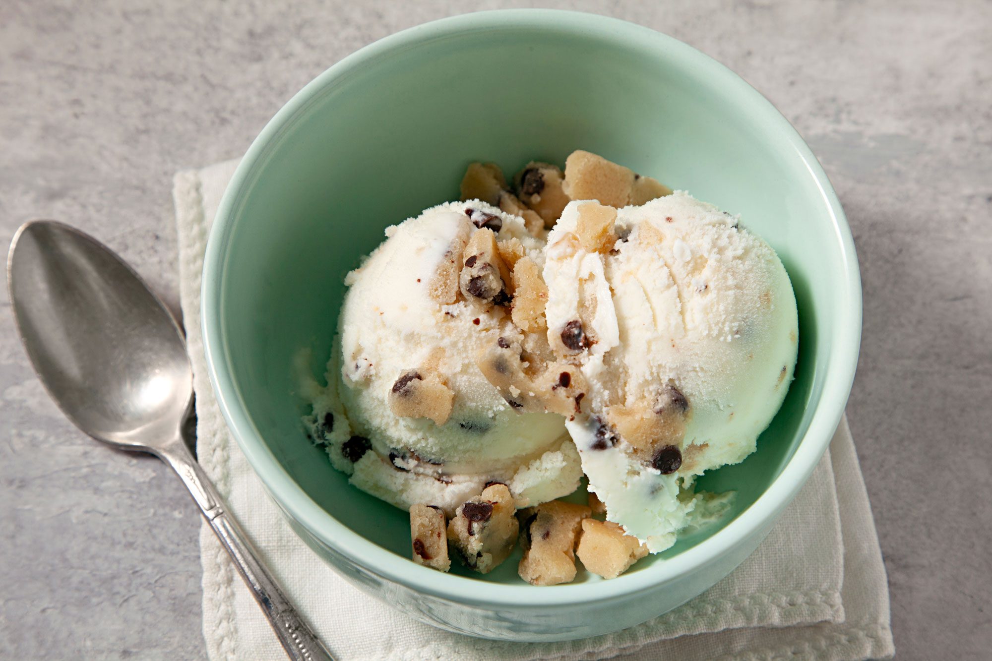 3/4th shot; grey background; Easy Cookie Dough Ice Cream served in a small green bowls over white kitchen towel with silver spoon;