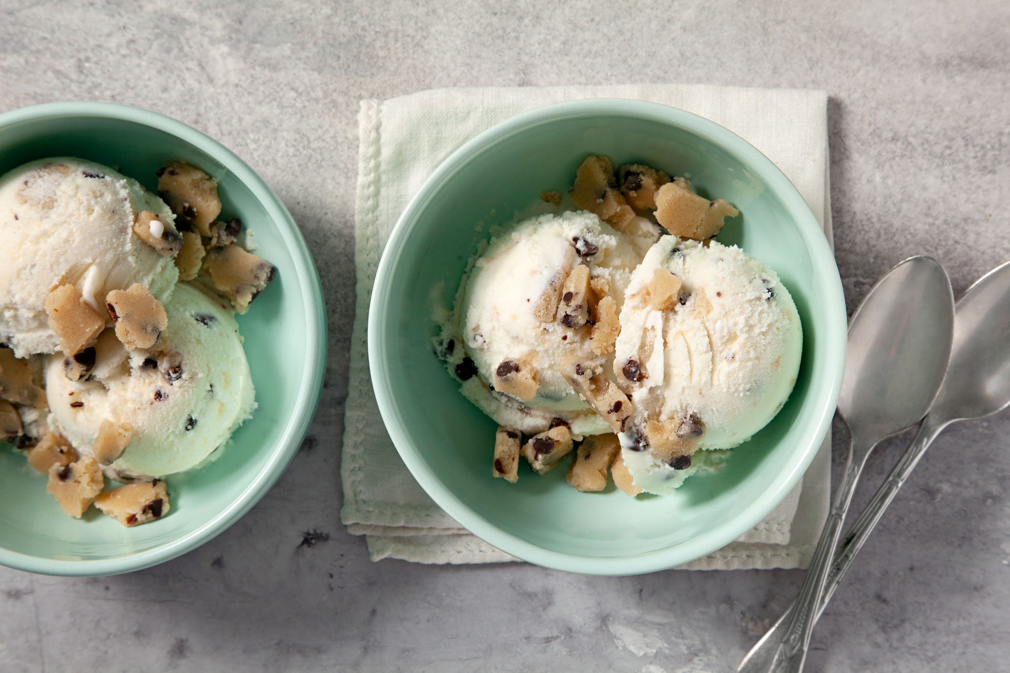 overhead shot; grey background; Easy Cookie Dough Ice Cream served in two small green bowls over white kitchen towel with two silver spoons;