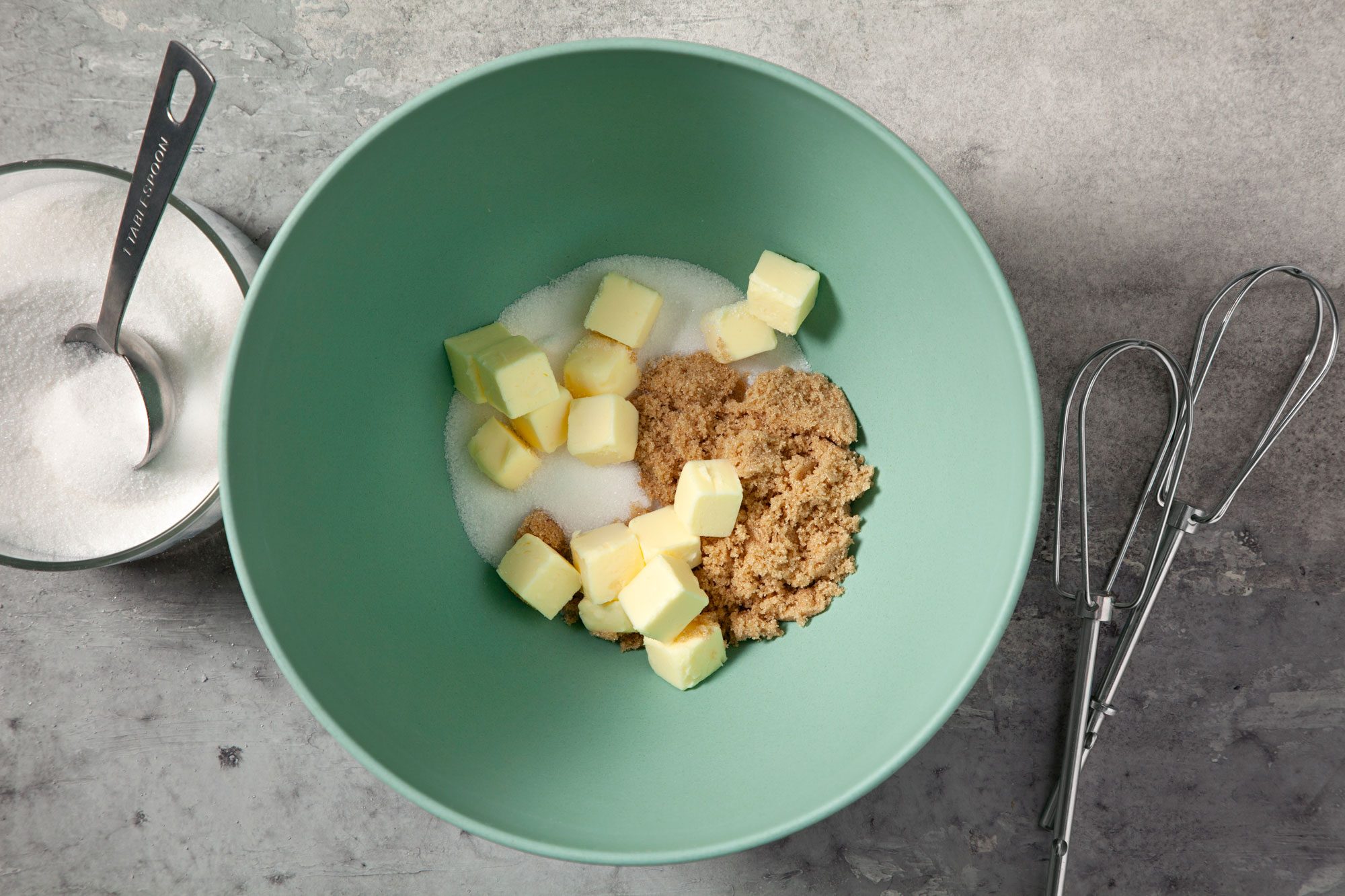 overhead shot; grey background; In a large bowl, butter and sugars; sugar in a cup with measuring spoon;