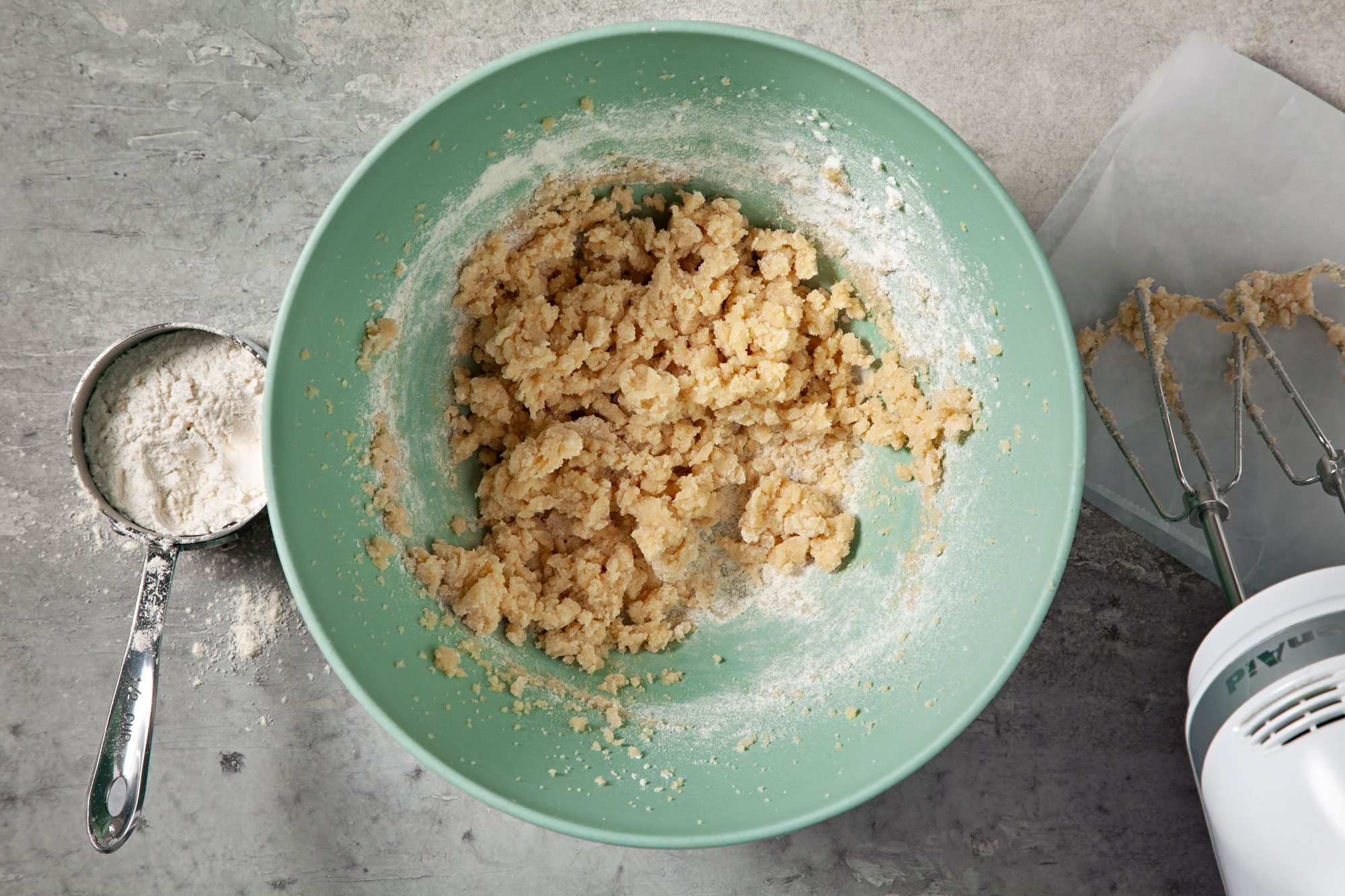 overhead shot; grey background; added flour into the mixture in a large bowl; flour in measuring cup; electric whisker;