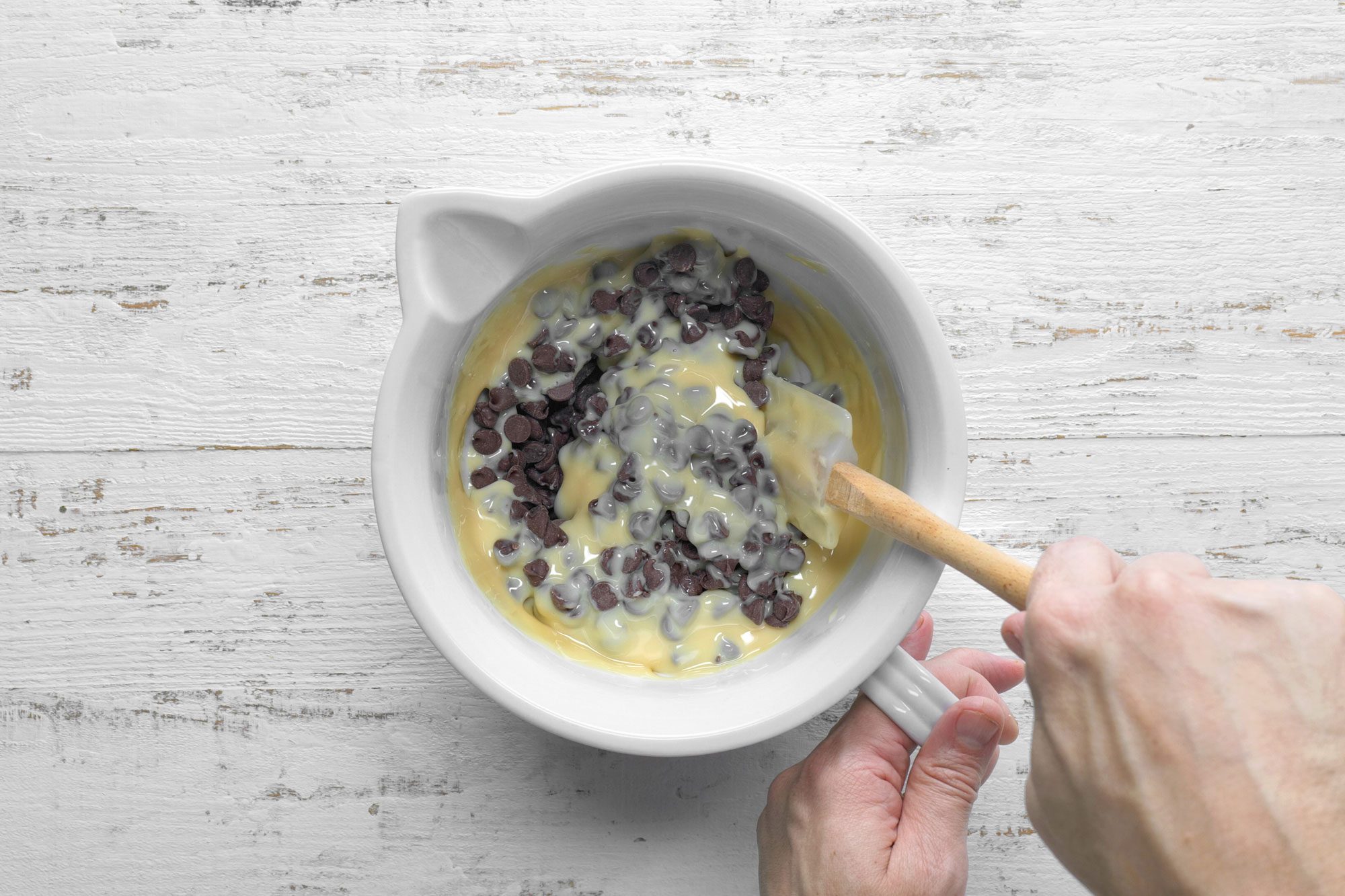 overhead shot; white wooden background; in a microwave-safe bowl, combining chocolate chips and milk and stiring with spatula;