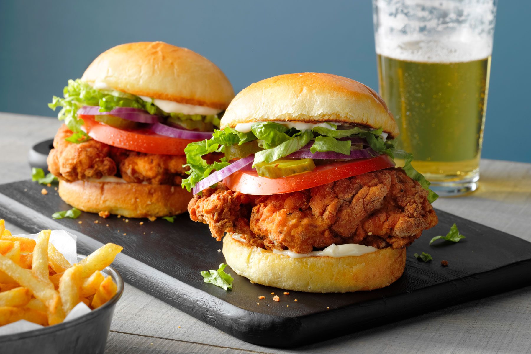 Two crispy fried chicken sandwiches are served on a black slate board. Next to them are a side of french fries in a small metal container and a glass of beer, set on a light gray table.