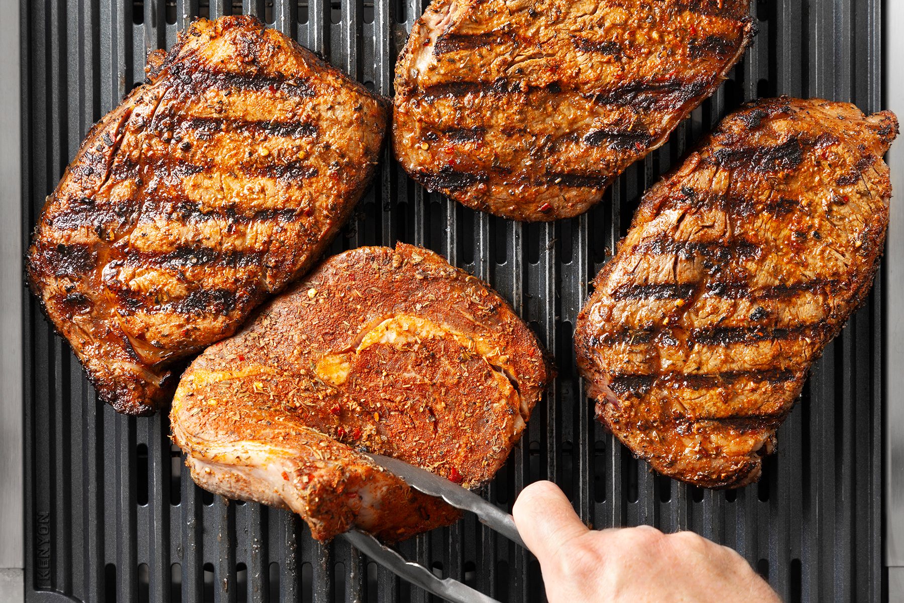 Four seasoned steaks are being grilled on a black grill. One of the steaks is being flipped with a pair of silver tongs, revealing grill marks. The steaks are perfectly browned, indicating they are well-cooked.