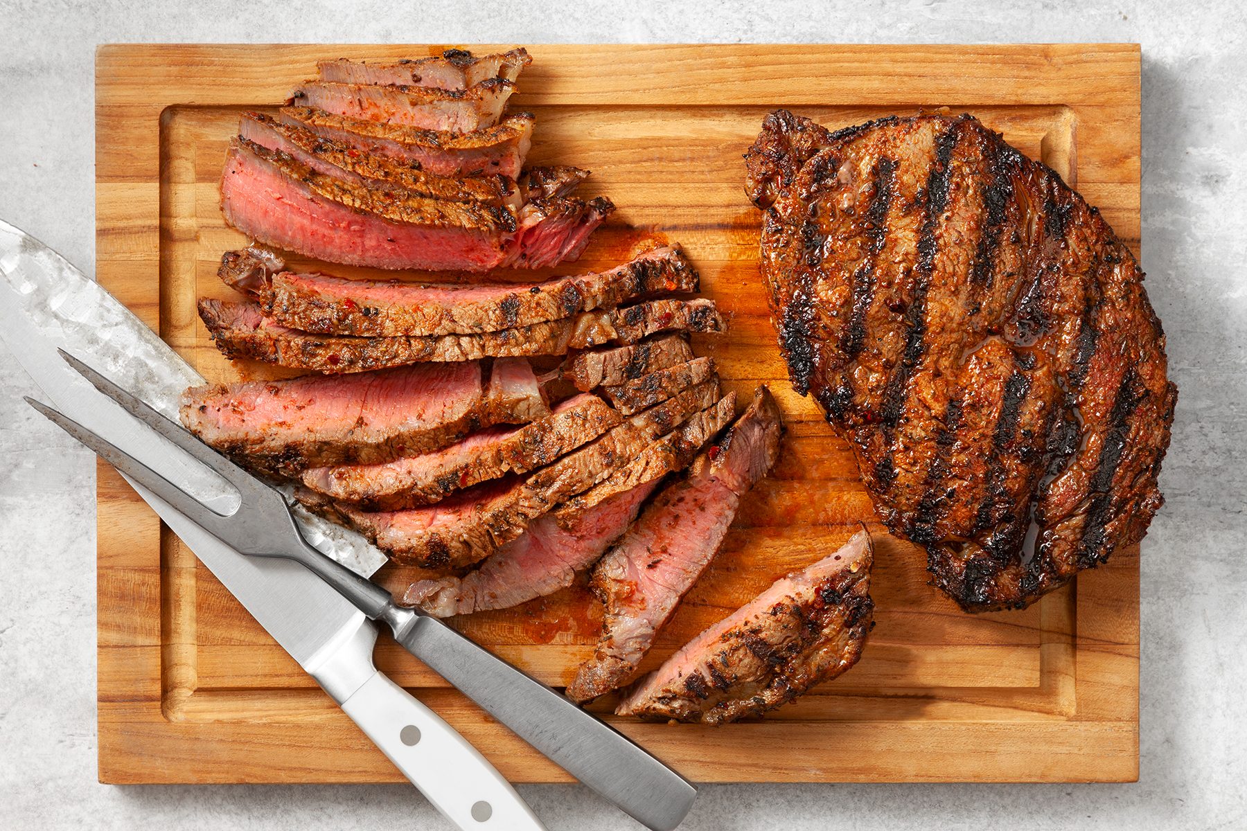 A wooden cutting board with a grilled steak, partially sliced. The steak has visible grill marks and is medium-rare in the center. 