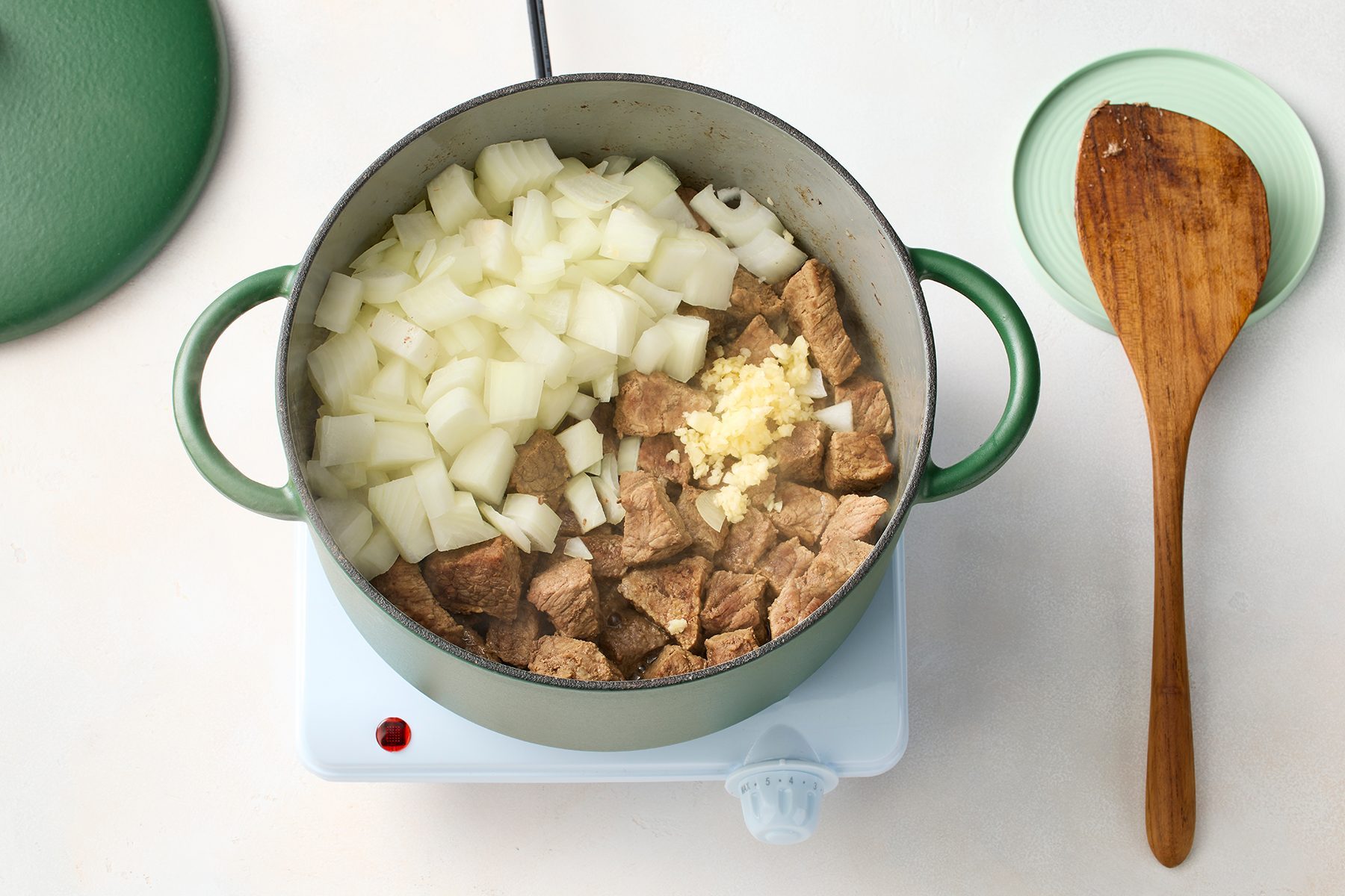 A green pot sits on a white electric stovetop, containing chopped onions, diced garlic, and browned meat. 