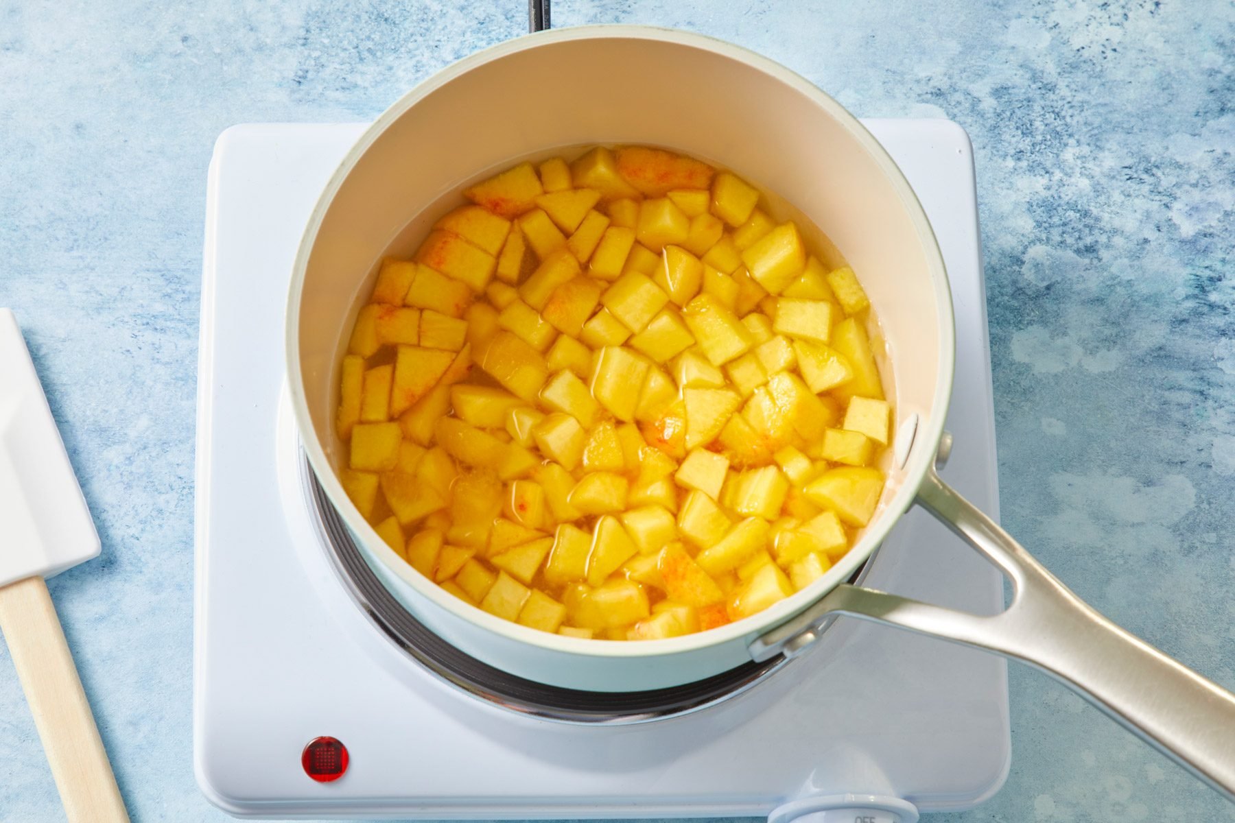 A white stovetop with a saucepan containing chopped peaches. A white spatula rests beside the stovetop on a blue, marbled countertop.