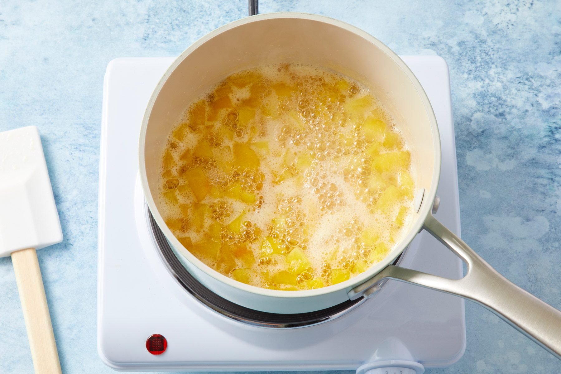 A white stovetop with a saucepan containing chopped peaches simmering in liquid. A white spatula rests beside the stovetop on a blue, marbled countertop.