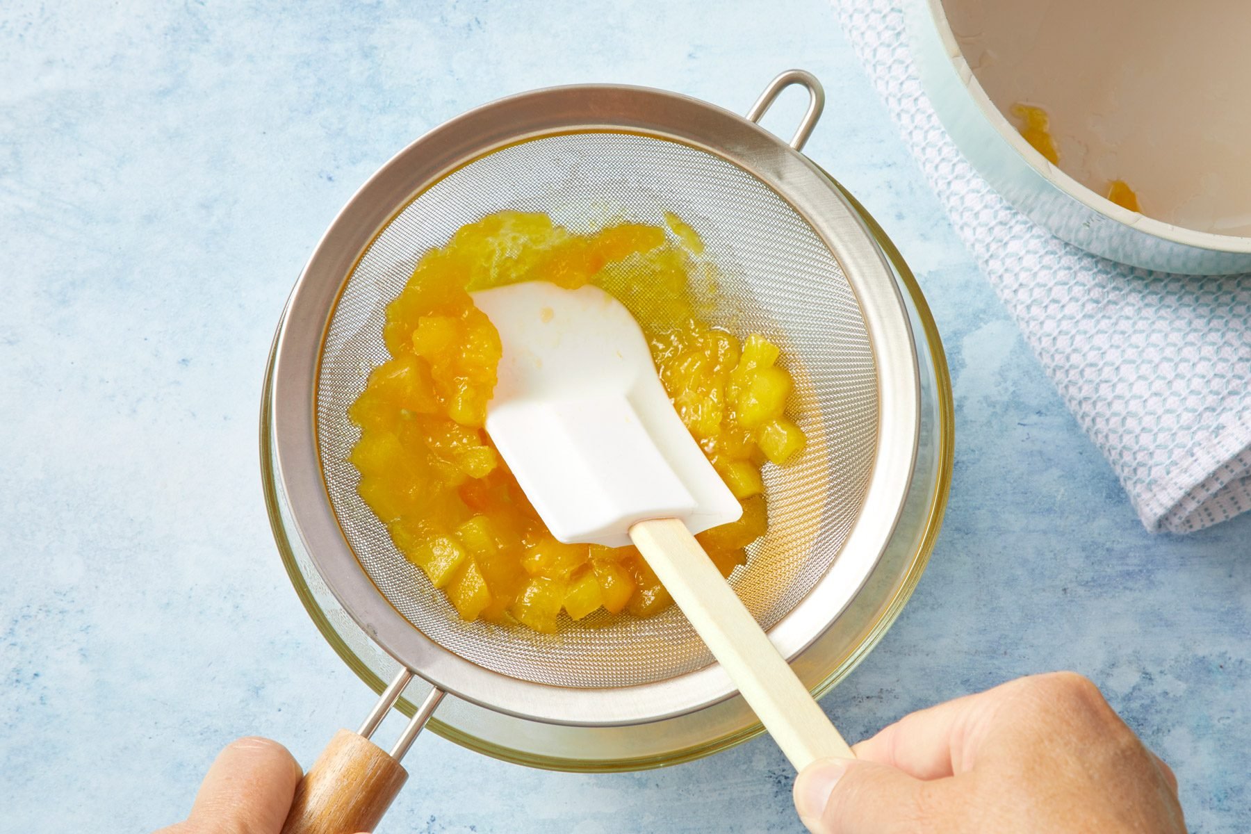 Hands using a white spatula to strain diced peaches through a metal sieve into a glass bowl. A towel and a bowl with remaining diced fruits are visible in the background on a light blue surface.