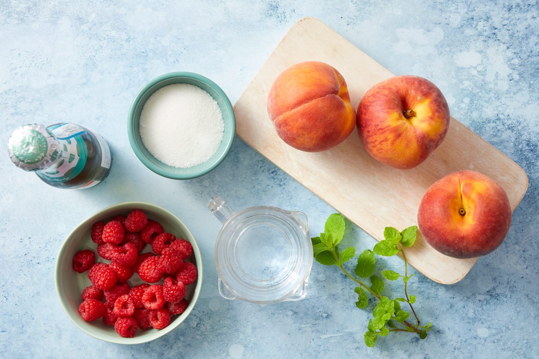 A flat lay of ingredients on a blue surface. There are three peaches on a cutting board, a bowl of raspberries, a clear bowl of sugar, a small glass jug of water, a bottle of champagne, and a sprig of fresh mint.