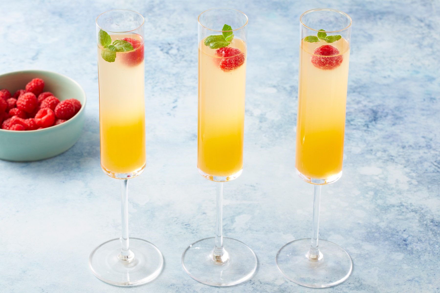 Three tall champagne flutes filled with a yellow-orange layered cocktail, garnished with raspberries and mint leaves, are arranged in a row against a blue background. A bowl of fresh raspberries is placed nearby.