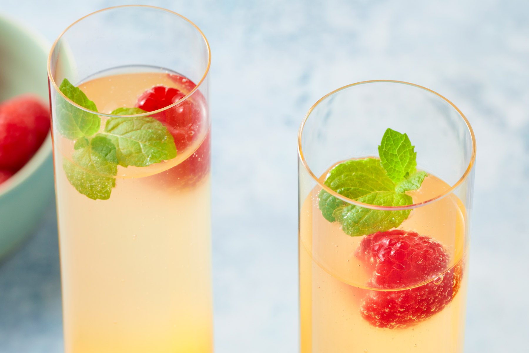 Two tall glasses filled with an orange beverage are garnished with mint leaves and raspberries. The glasses are set against a light blue background, with a bowl of raspberries partially visible in the corner.