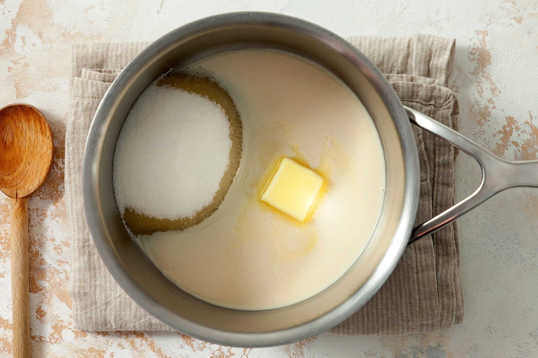 overhead shot; white textured background; combined sugar, milk and butter in a saucepan placed over kitchen towel with long wooden spoon;