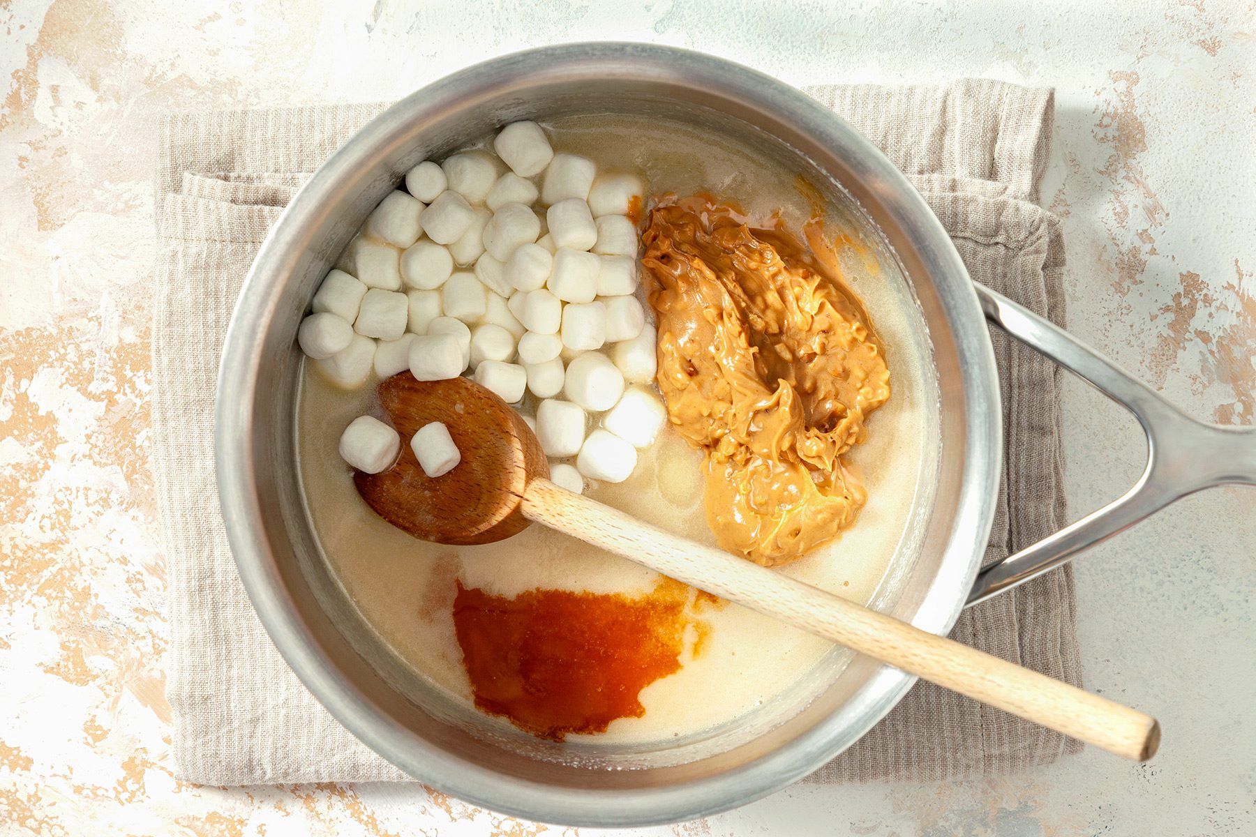 overhead shot; white textured background; added peanut butter, marshmallows and vanilla in saucepan placed over kitchen towel;