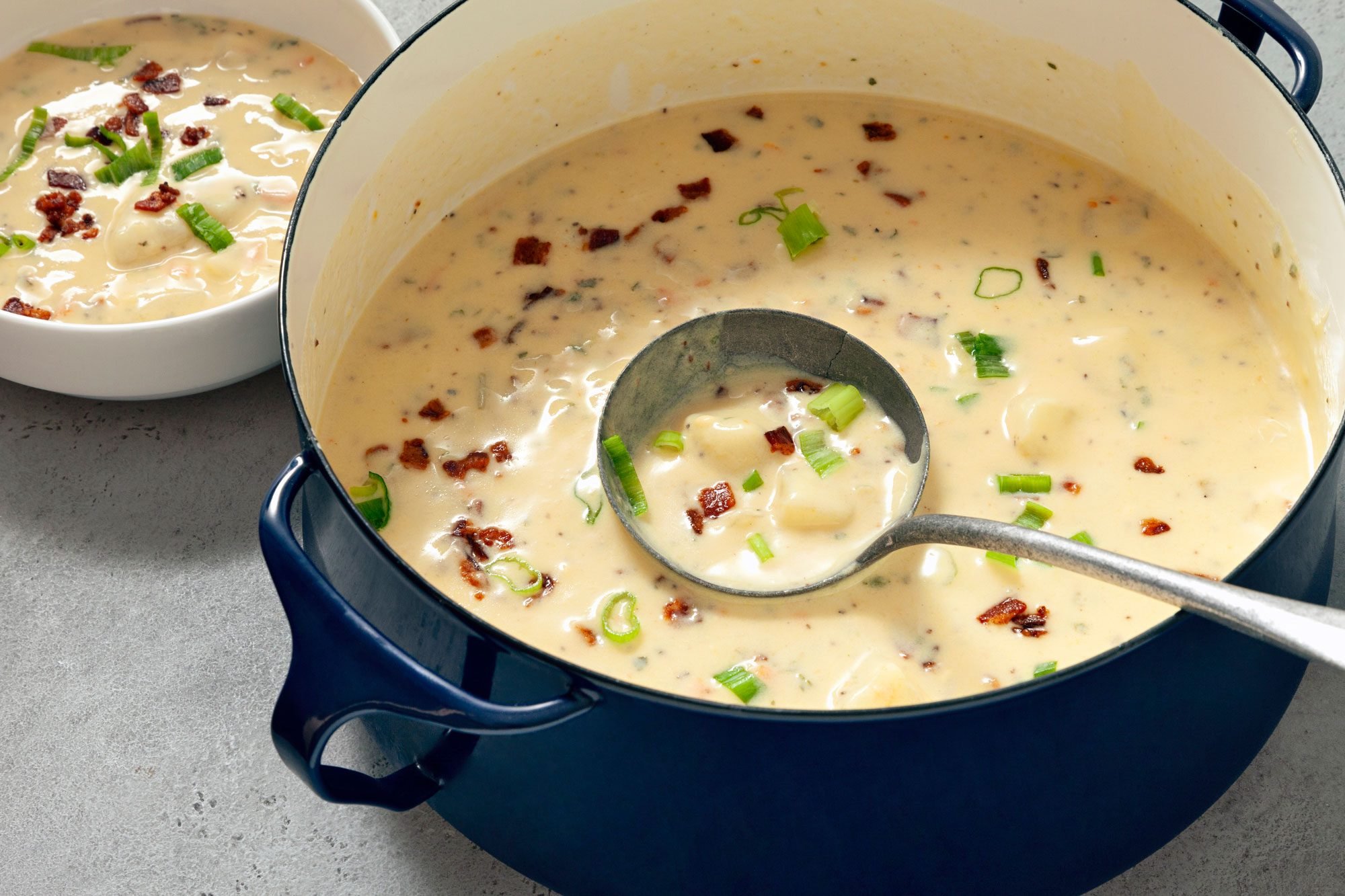 3/4th shot; horizontal; light grey background; Potato Soup in a large skillet with long silver serving spoon in it; Potato Soup in small white bowl;