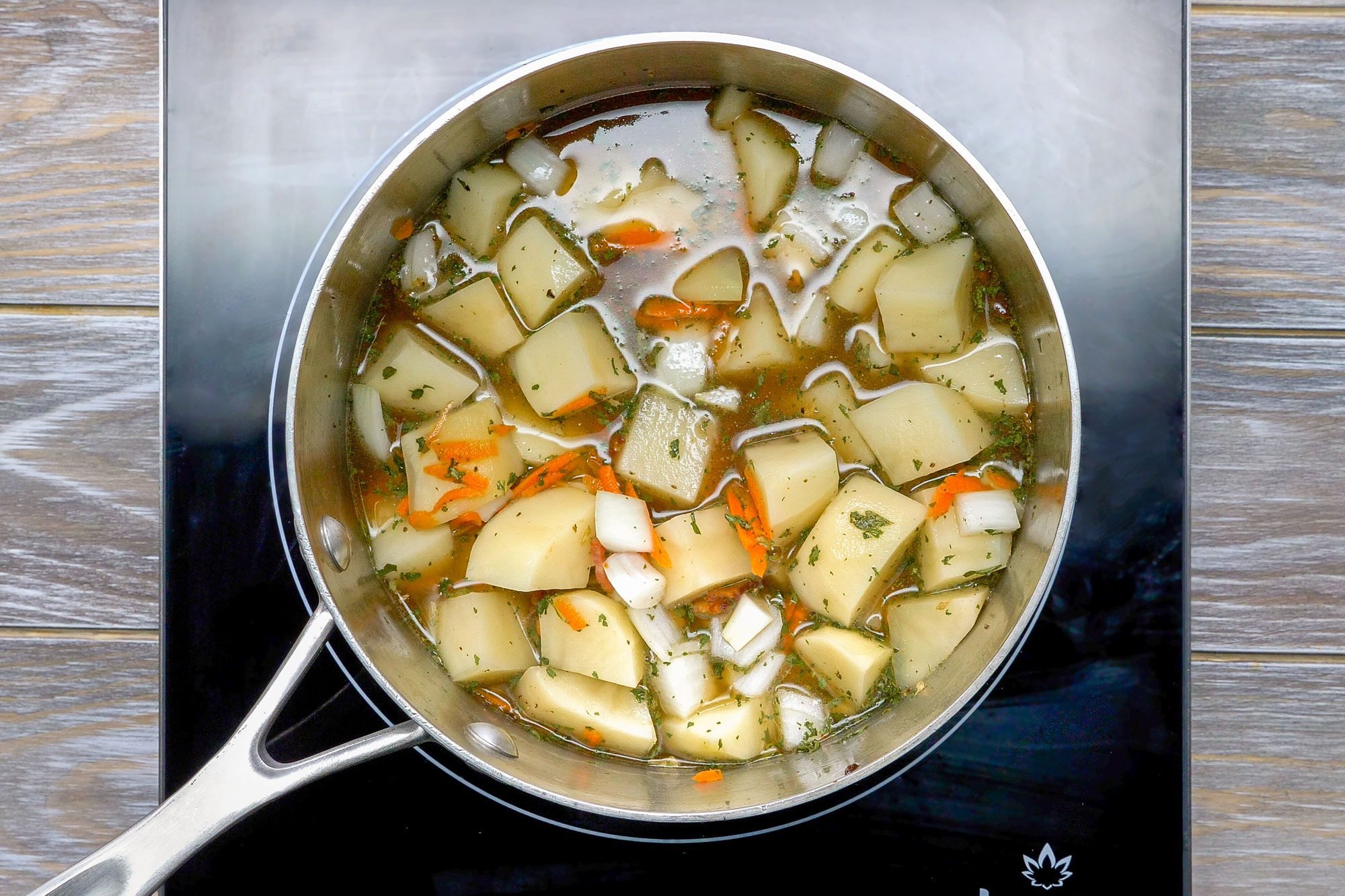 Overhead shot of same saucepan; add vegetables; seasonings and broth; bring to boil; simmer; covered; until potatoes are tender; induction; wooden background;