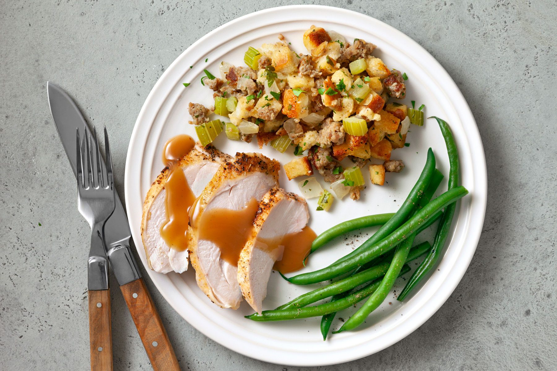 A white plate with a meal consisting of sliced roasted turkey breast covered in gravy, stuffing with vegetables, and green beans.