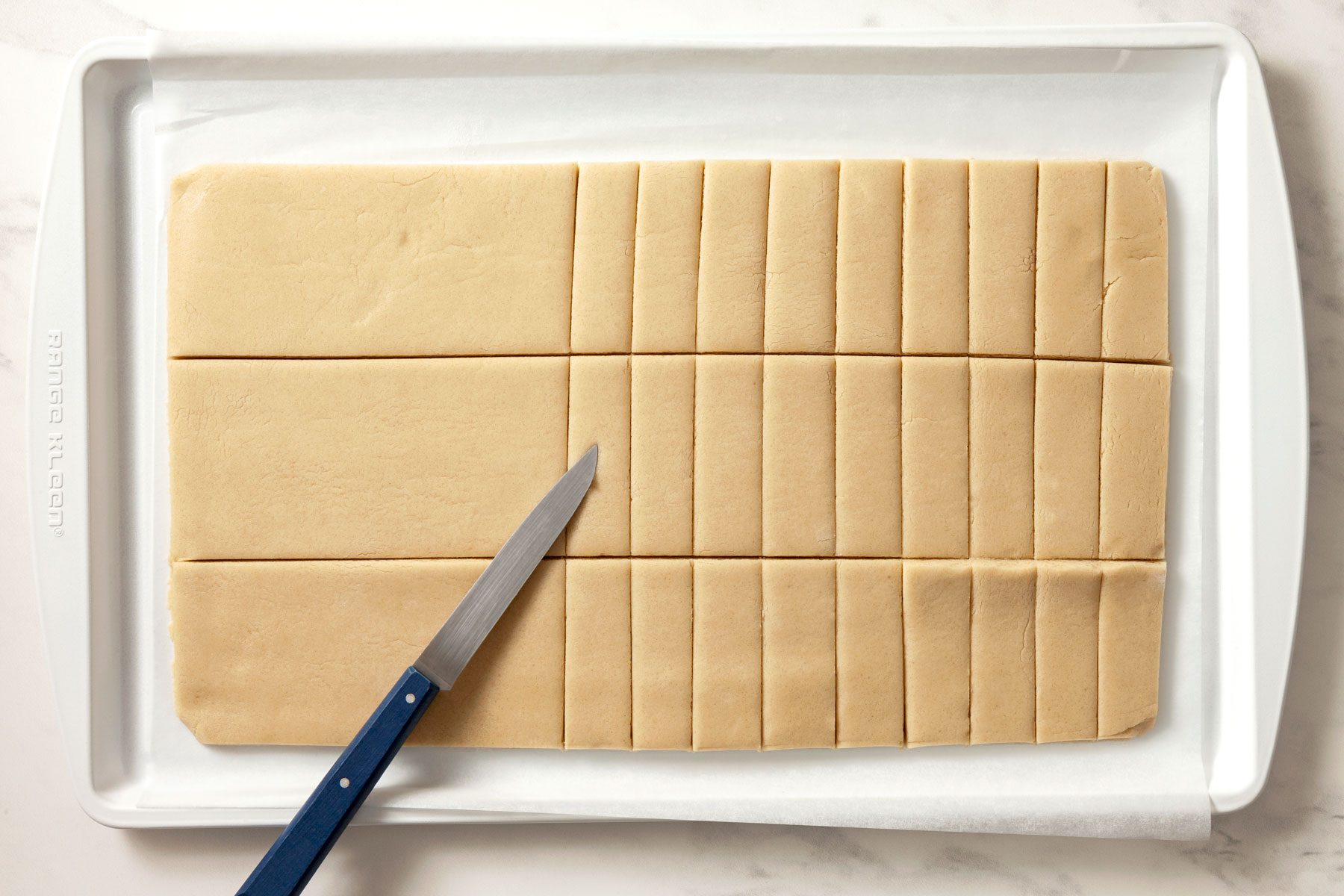 A slab of dough on a baking sheet is cut into strips. A knife rests on the slab.