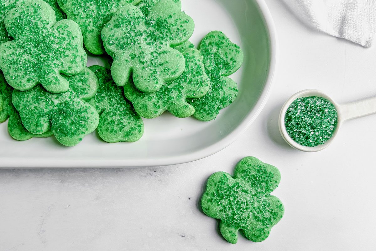 Taste of Home Shamrock cookies on a white ceramic plate next to a white ceramic spoon filled with green sugar on a marble surface