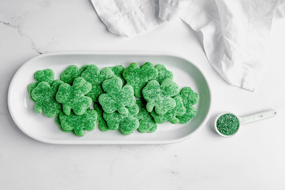 Taste of Home Shamrock cookies on a white ceramic plate next to a white ceramic spoon filled with green sugar on a marble surface.