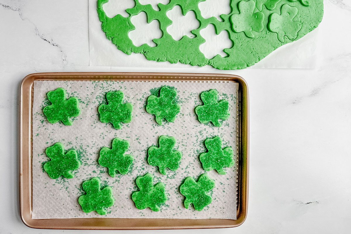 Taste of Home Shamrock cookie dough cut out on a parchment sheet next to a parchment lined baking sheet on a marble surface.