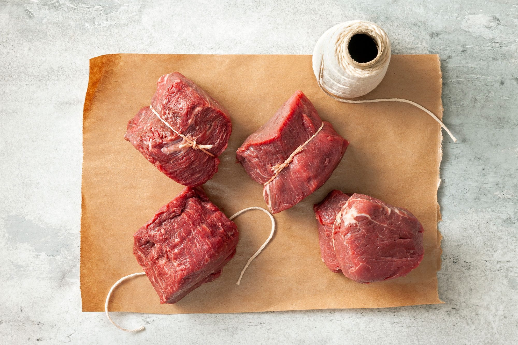 overhead shot; grey textured background; Using kitchen string, tied a loop around each tenderloin steak and placed over kitchen brown paper; string roll