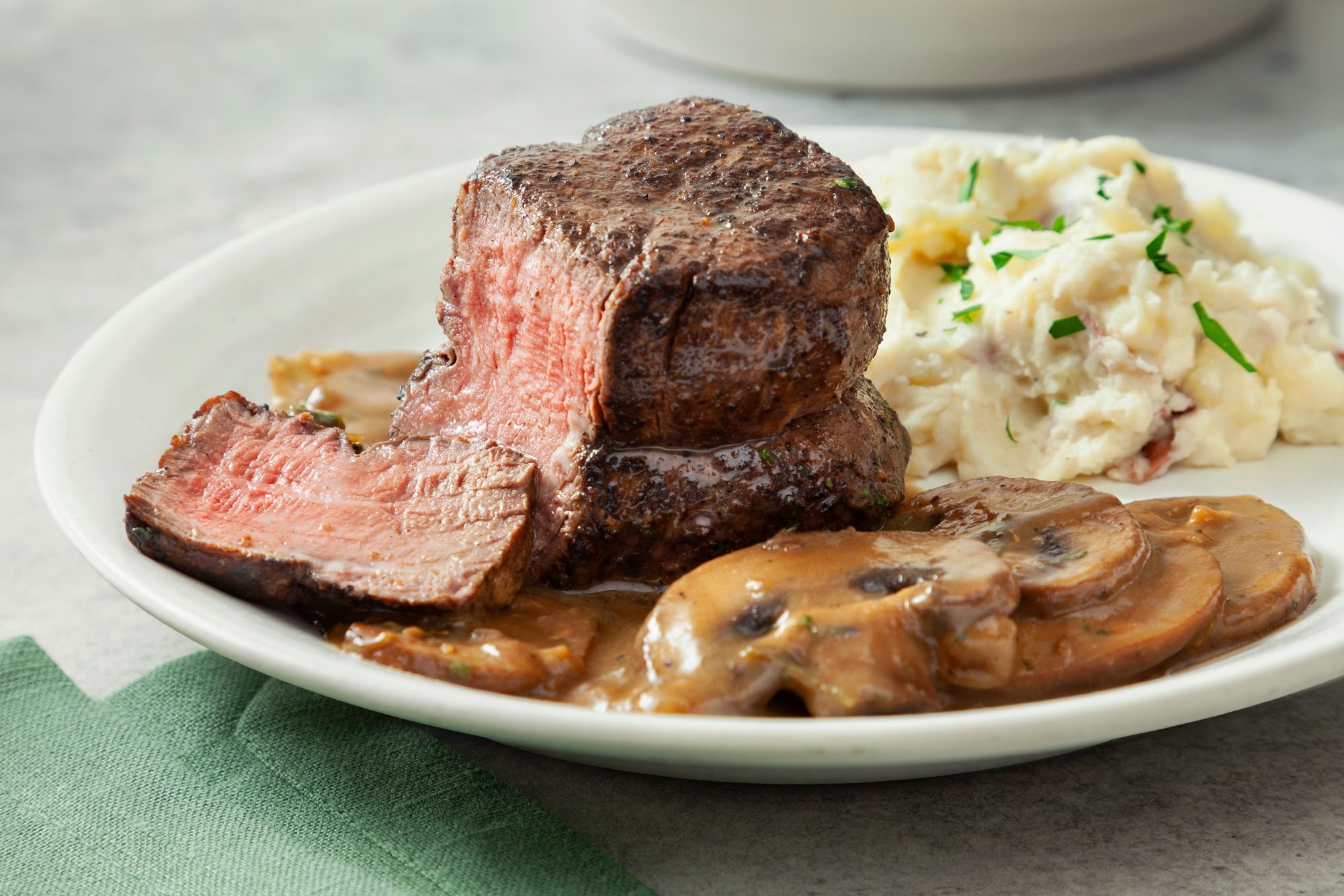 3/4th shot; grey background; Copycat Cheesecake Factory Steak Diane's slice cut from side portion and placed in small white plate served with mashed potatoes with green kitchen napkin;