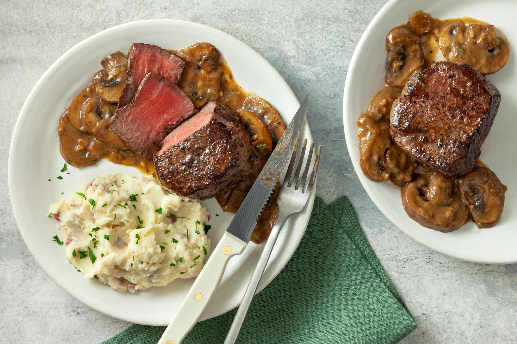 overhead shot; grey background; Copycat Cheesecake Factory Steak Diane in two small white plates served with mashed potatoes with green kitchen napkin and sliver fork and spoon;