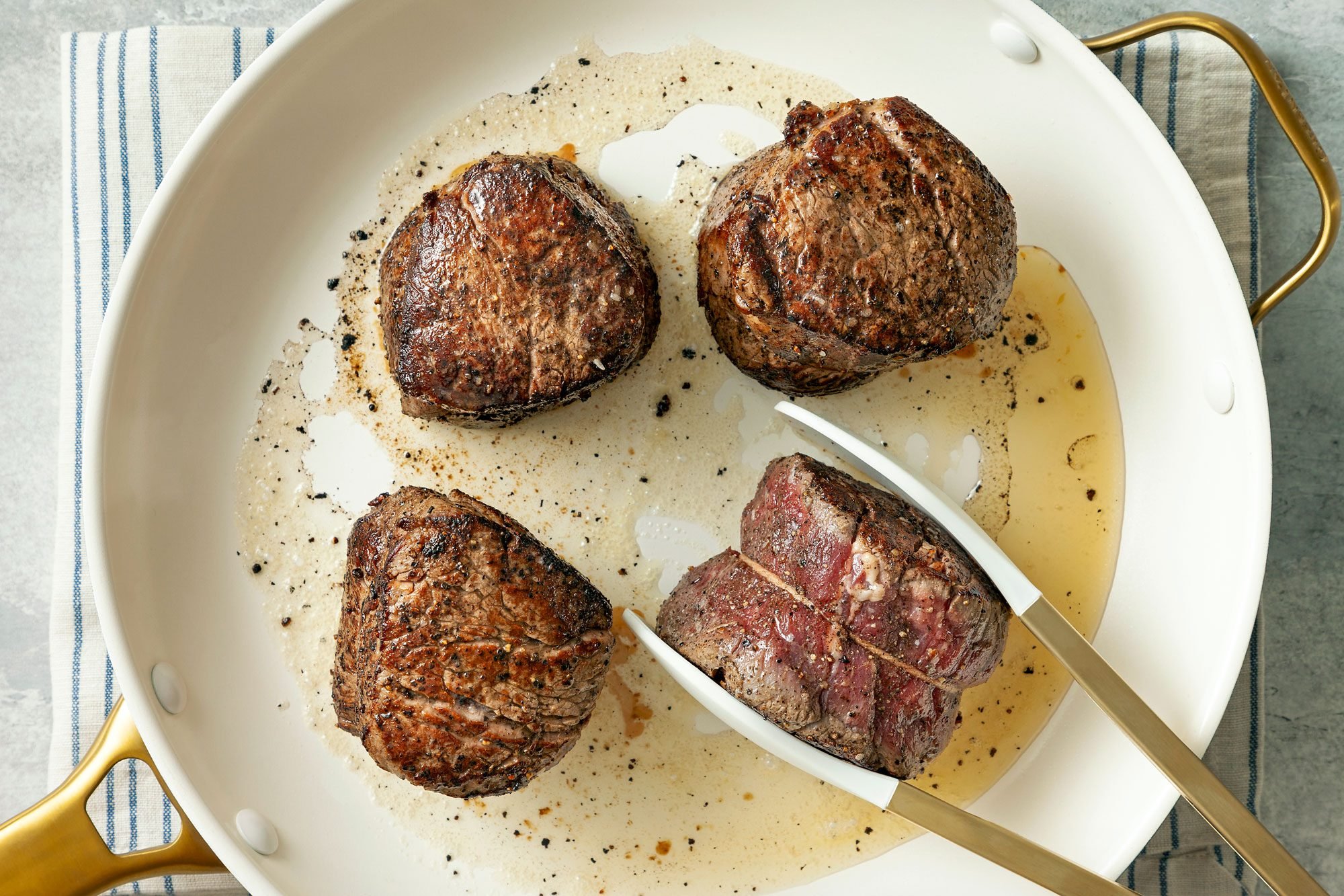 overhead shot; grey textured background; In a large skillet, heating oil over medium heat, Added steaks; cooking until meat reaches desired doneness; steak in tongs; pan placed over kitchn towel;