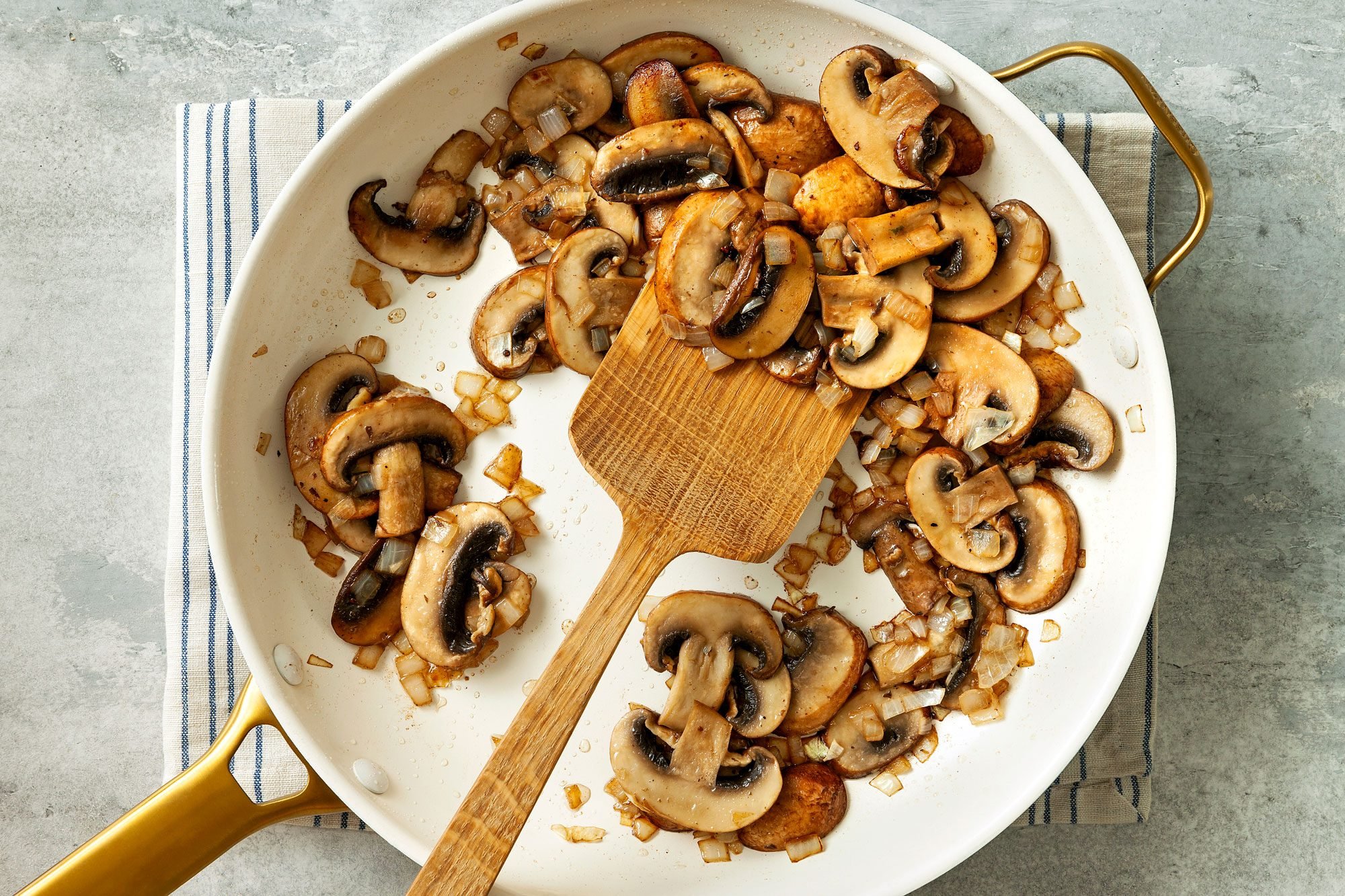 overhead shot; grey textured background; Added mushrooms and onion to skillet; cooked and stiring with wooden spatula, Add garlic; pan placed over kitchen towel;