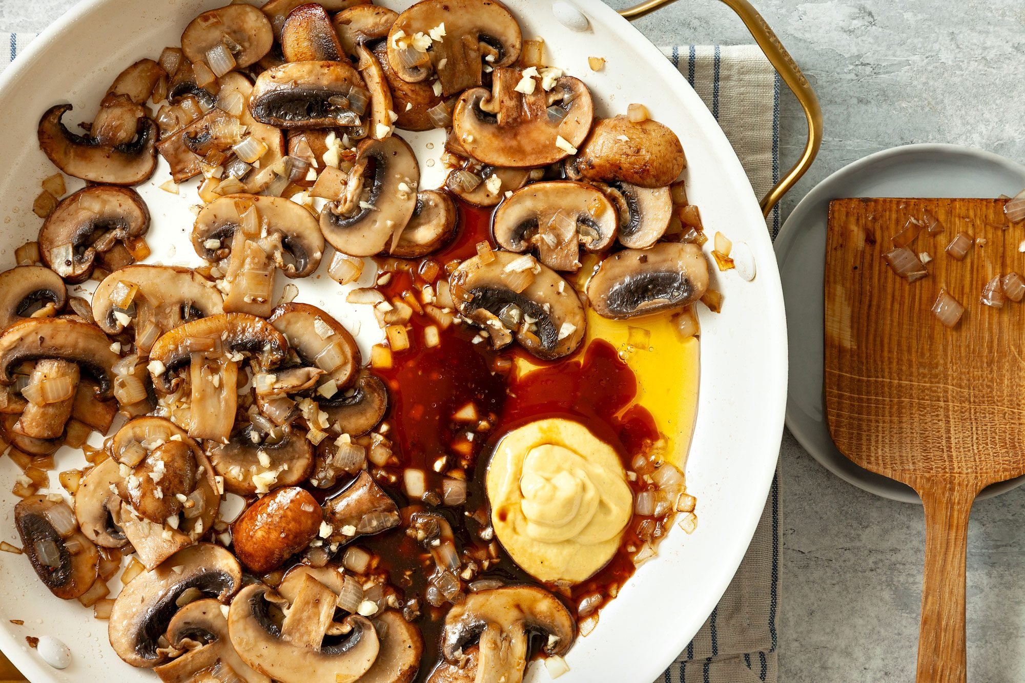 overhead shot; grey textured background; Stir in Worcestershire, Dijon and honey over mushrooms in pan placed over kitchen towel; wooden spatula placed over a small plate on right side;