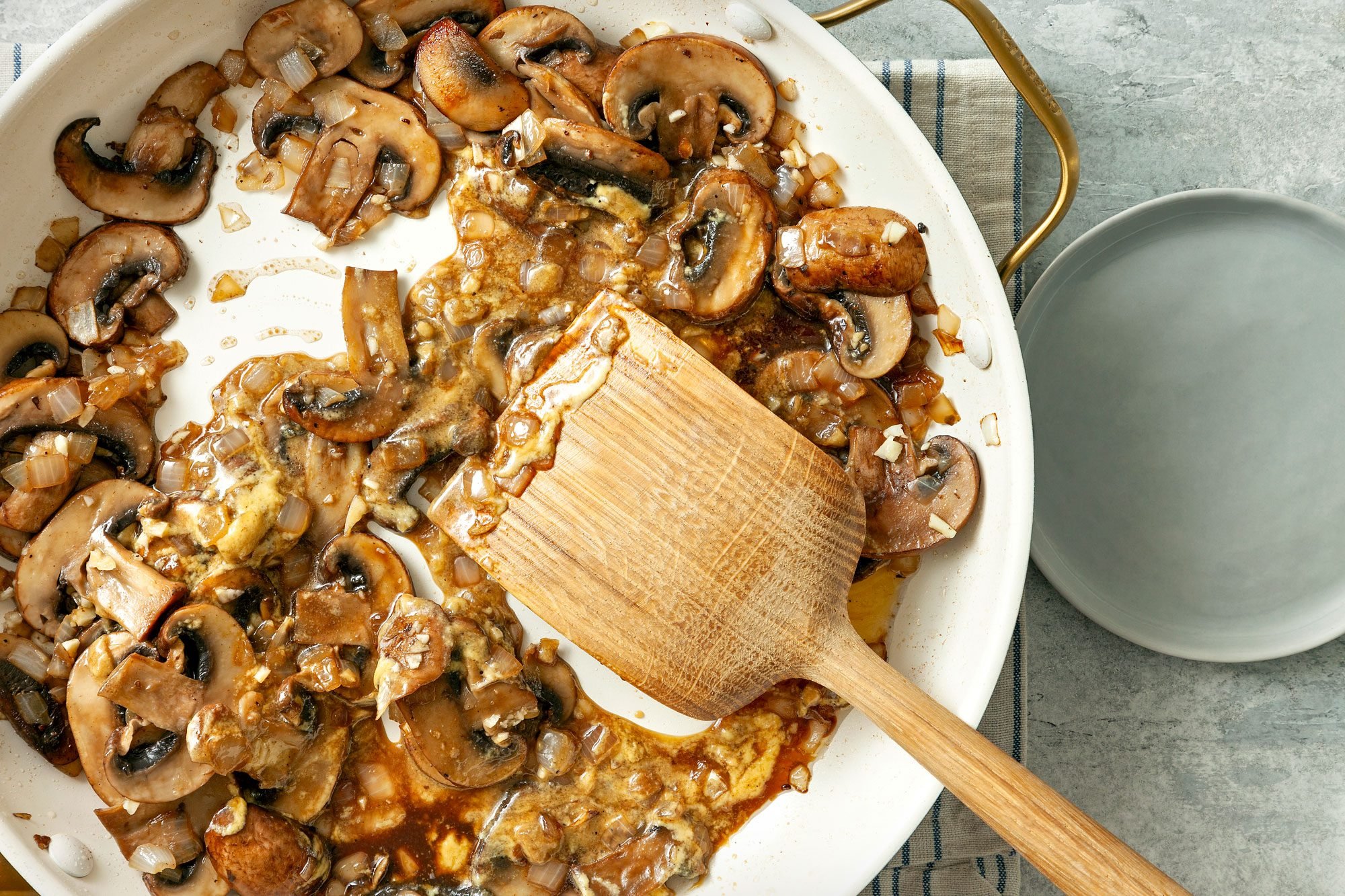 overhead shot; grey textured background stiring mushrooms in pan with wooden spatula; placed over kitchen towel; grey small plate placed on right side;