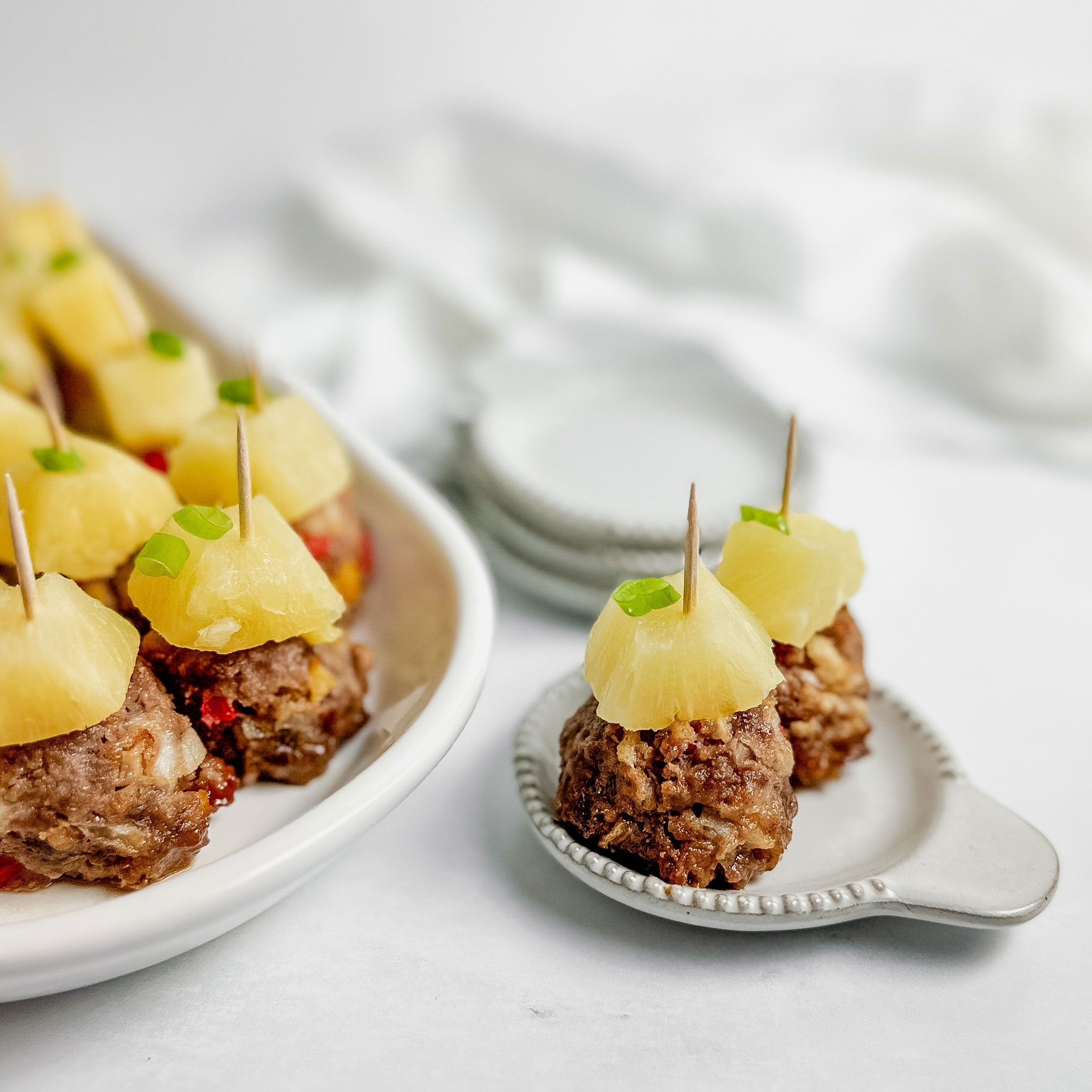 Taste of Home teriyaki meatballs topped with pineapple chunks in a white ceramic tray next to a linen napkin and mini plates on a marble surface.