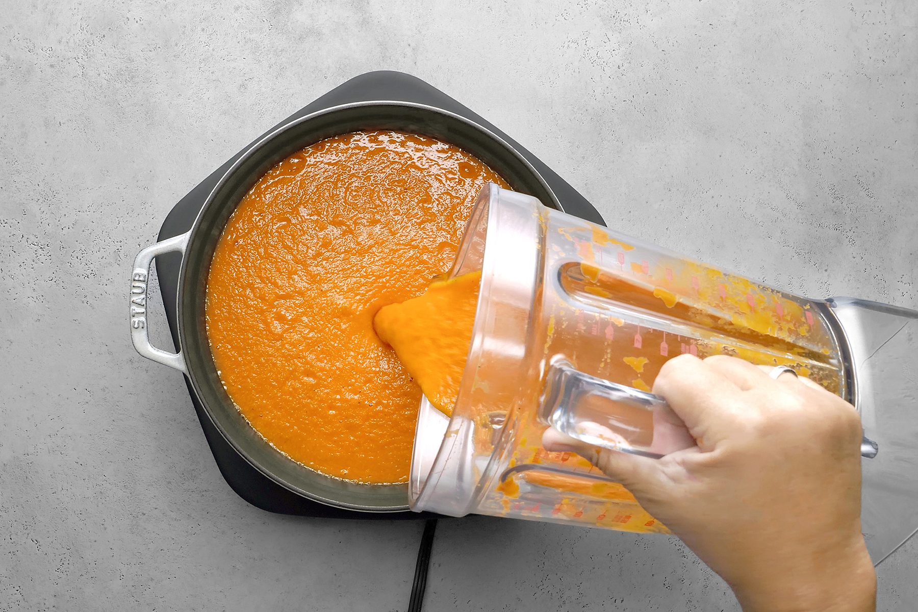 A person pours a thick, orange soup from a blender into a pot on a stovetop. 