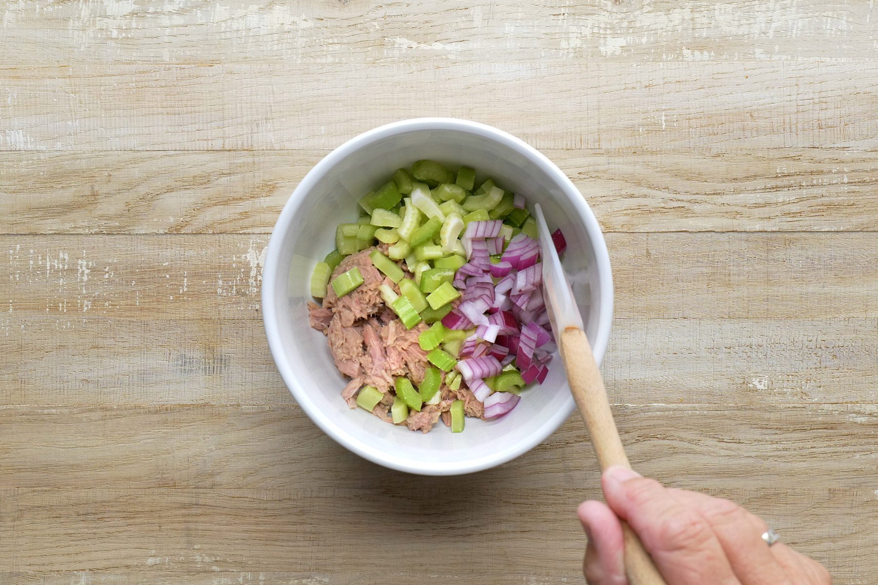 A person stirring a bowl containing chopped celery, red onion, and chunks of tuna with a wooden spoon, on a wooden surface background.