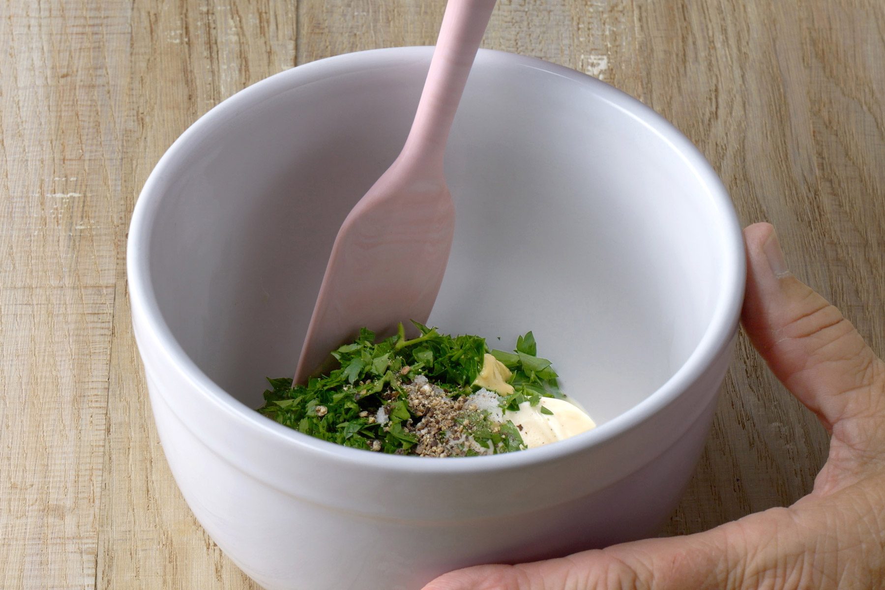 A hand holds a white bowl with chopped herbs, mayonnaise, and spices being mixed with a spatula on a wooden surface.