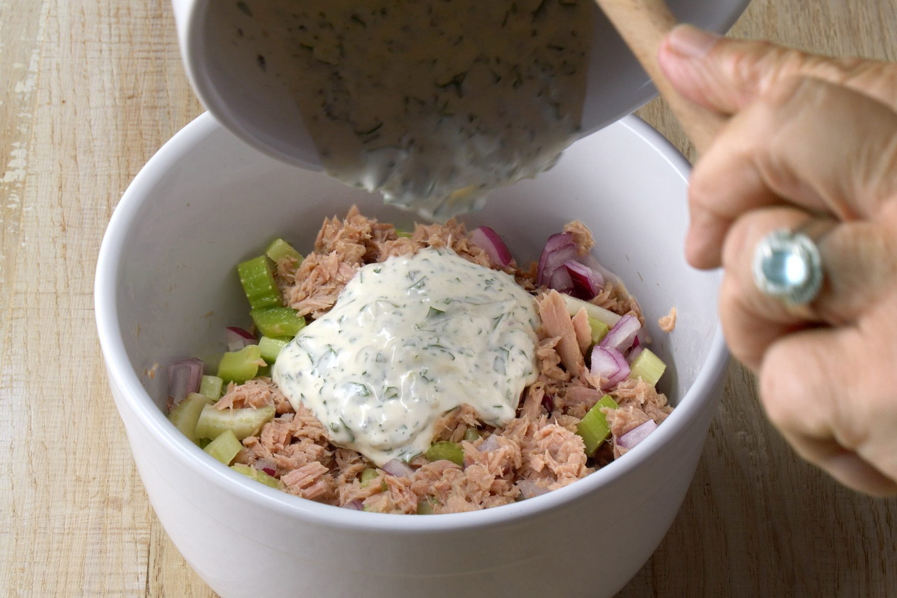 A person pouring mayonnaise over tuna mixture in a white bowl