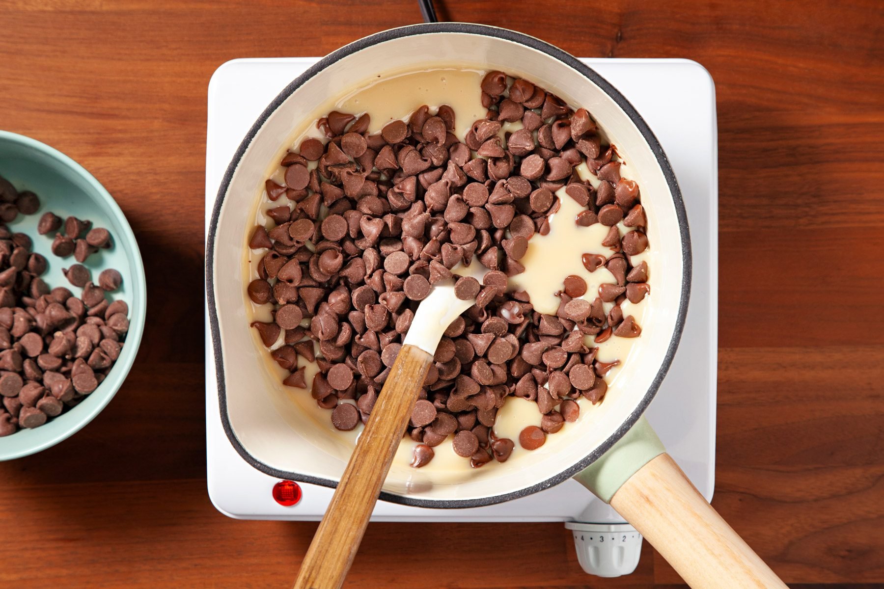 A saucepan filled with chocolate chips and sweetened condensed milk is on a stovetop. A spatula is stirring the mixture. A bowl with additional chocolate chips is to the side on the wooden counter.