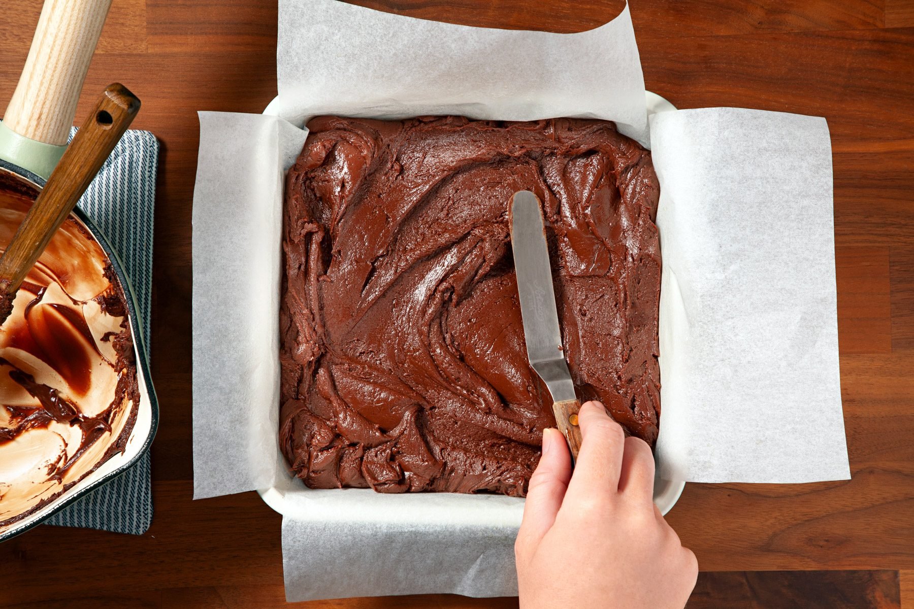 Fudge mixture is in a baking pan lined with paper. A hand can be seen using the back of a spatula to smooth the mixture evenly into the pan. A saucepan with leftover fudge mix is on the side on the wooden surface.