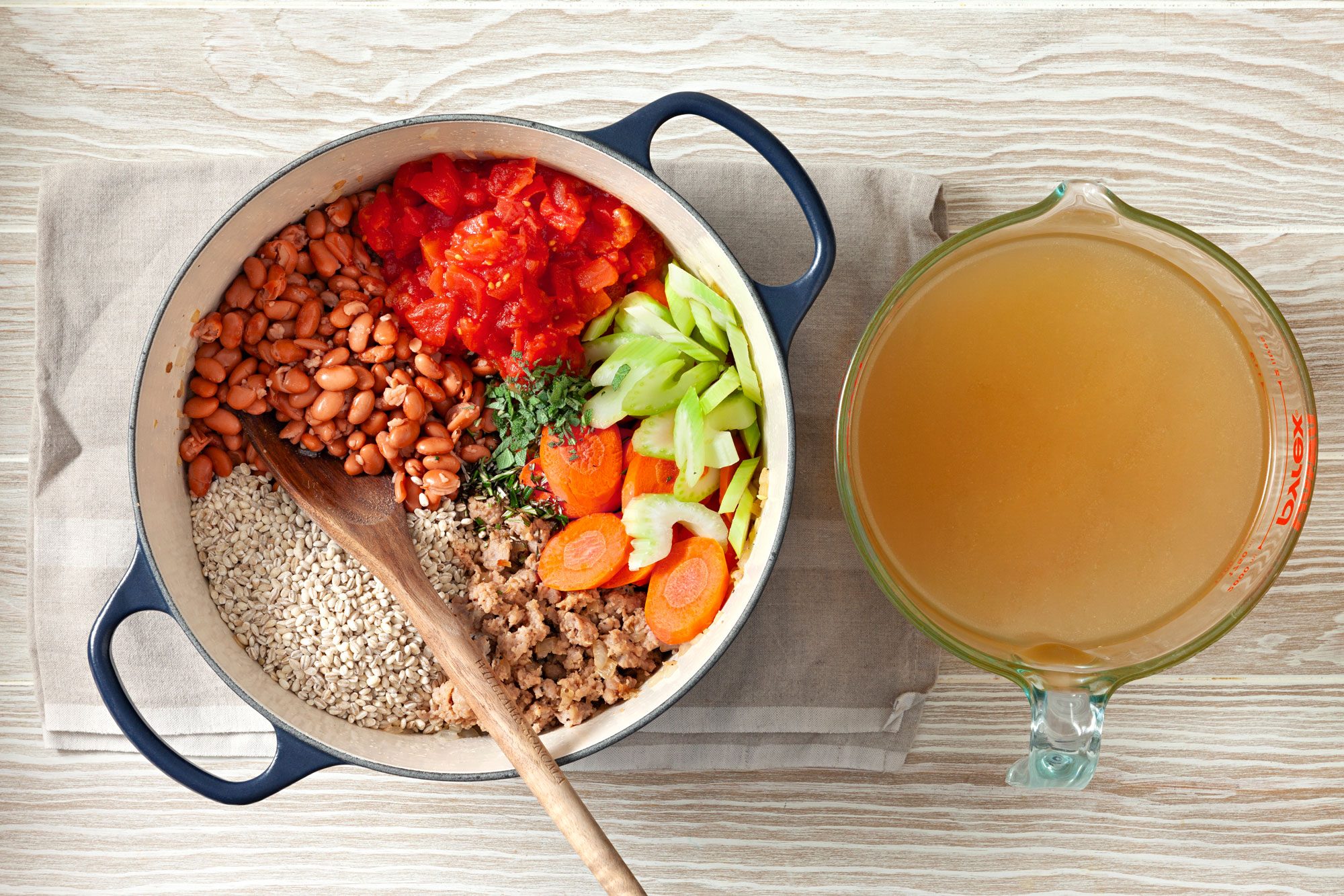 The broth in a jar, beans, tomatoes, barley, carrot, celery, sage and rosemary in dutch oven with wooden spoon