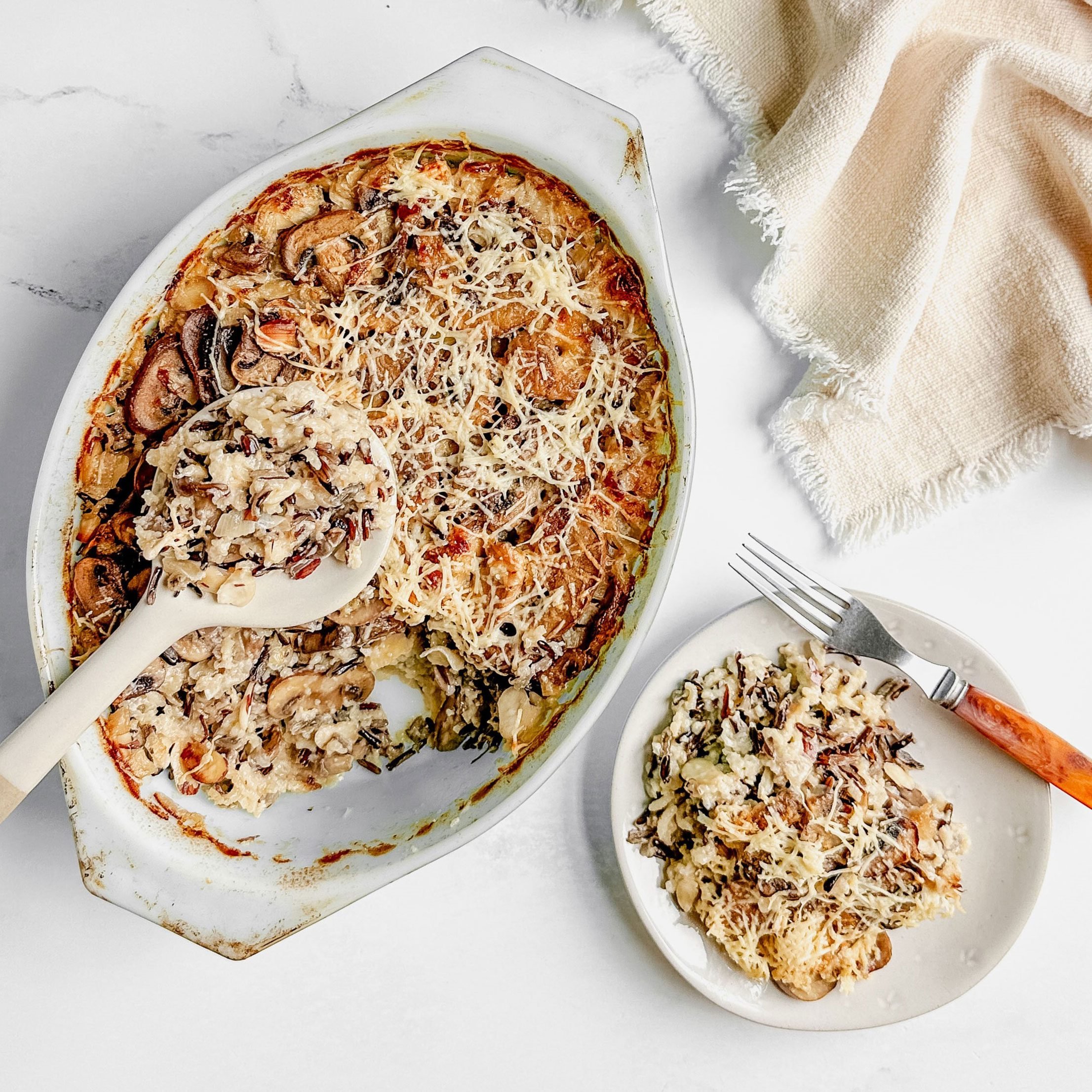 Taste of Home wild rice casserole in a pyrex dish and a ceramic plate next to a linen napkin on a marble surface.