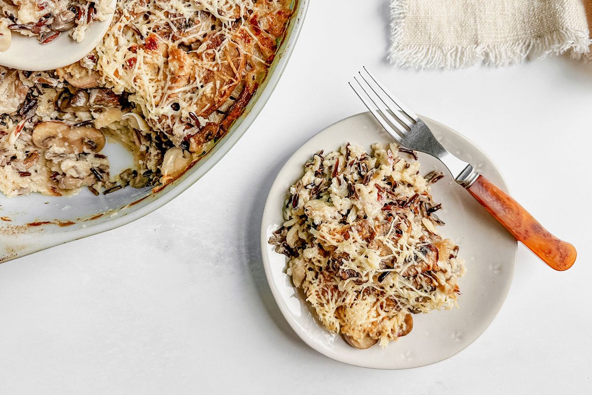 Taste of Home wild rice casserole in a pyrex dish and a ceramic plate next to a linen napkin on a marble surface.