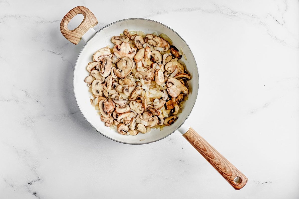 Mushrooms and onions for Taste of Home wild rice casserole in a skillet on a marble surface.