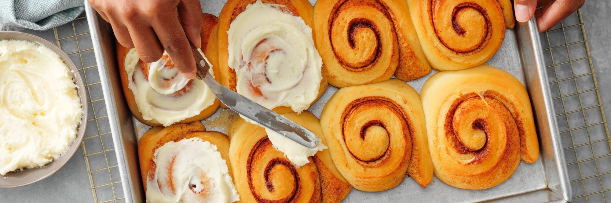 close up of hands frosting freshly baked cinnamon rolls